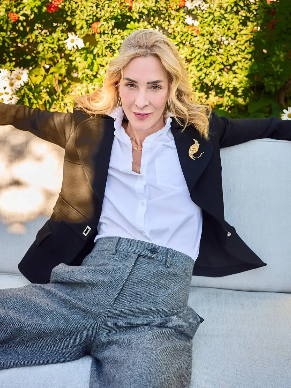 Portrait of an executive woman with blonde hair sitting on a light-colored outdoor sofa, she is wearing a black blazer with a gold pin, a white shirt.