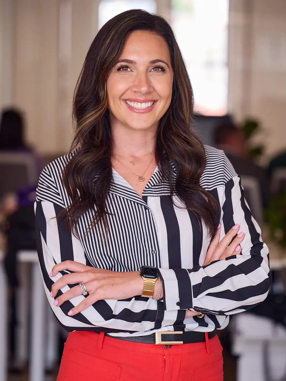 A smiling woman with long dark hair, wearing a black and white striped blouse, red pants, and a gold watch, photographed standing with arms crossed in an office setting.