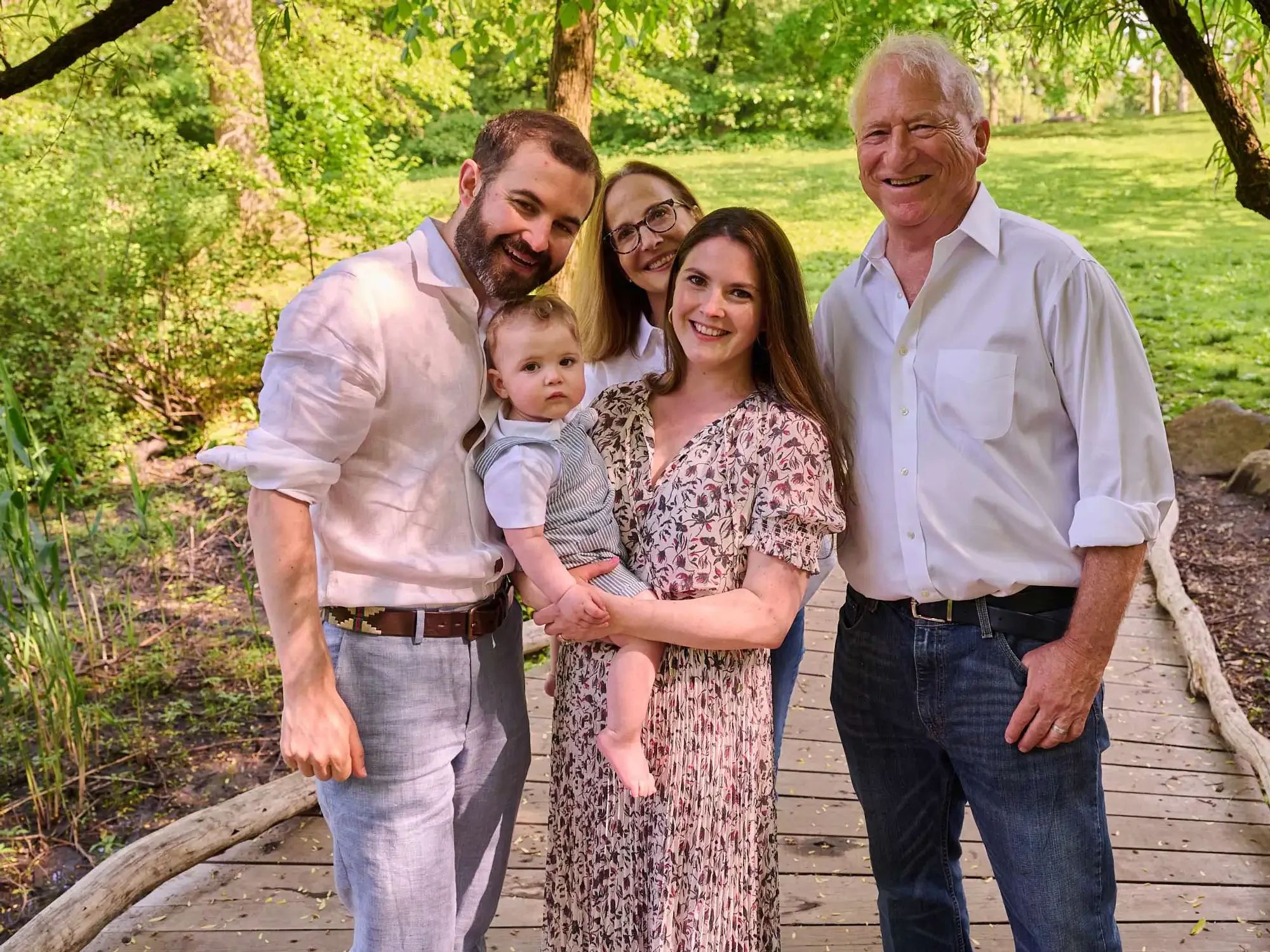 Family photograph of a multigenerational family group photo outdoors in a park, with five adults and a toddler, standing on a wooden path surrounded by green trees.