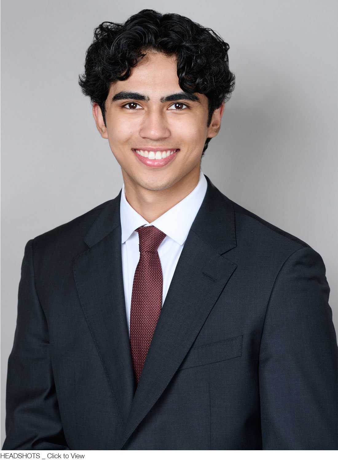 A corporate headshot of a young man with black curly hair, wearing a black suit, white shirt, and a maroon tie, smiling at the camera against a grey background.