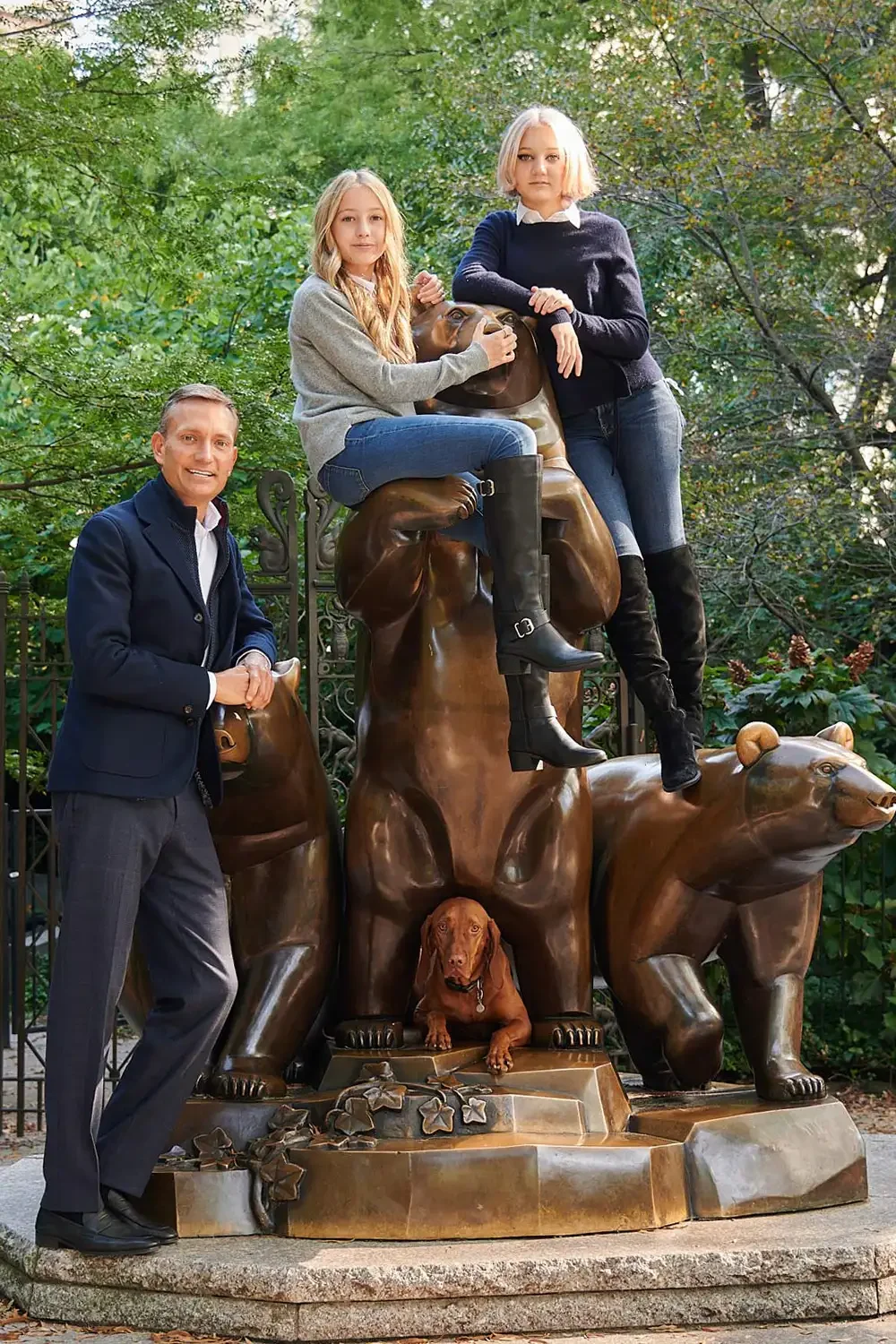 Photo of three people and a dog in front of a bronze sculpture of bears and a dog. Two women are sitting on top of the sculpture, one man is standing beside it, and a real dog is lying at the base.