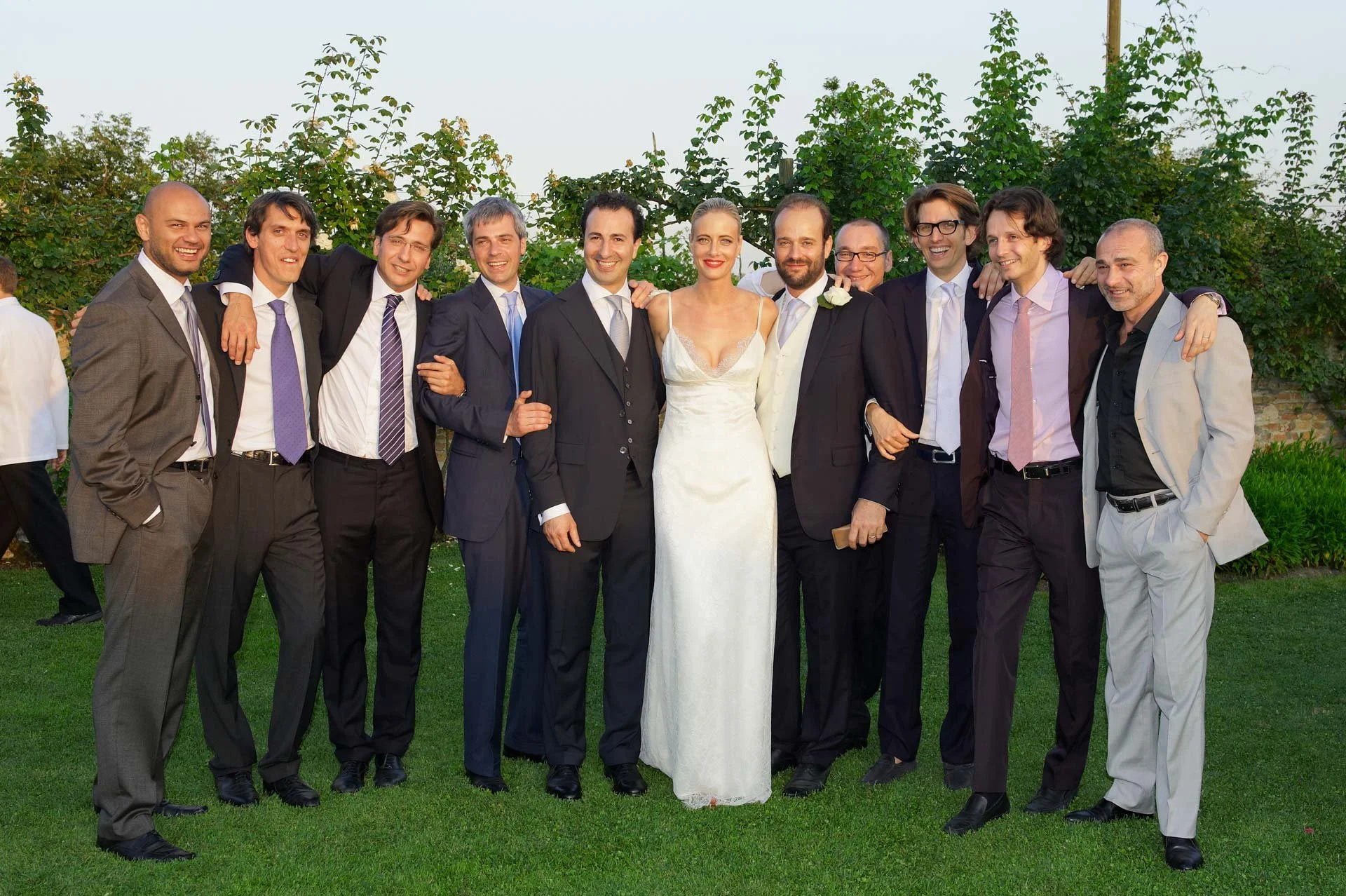 Group of people in formal attire standing outdoors during a wedding celebration, with greenery in the background.