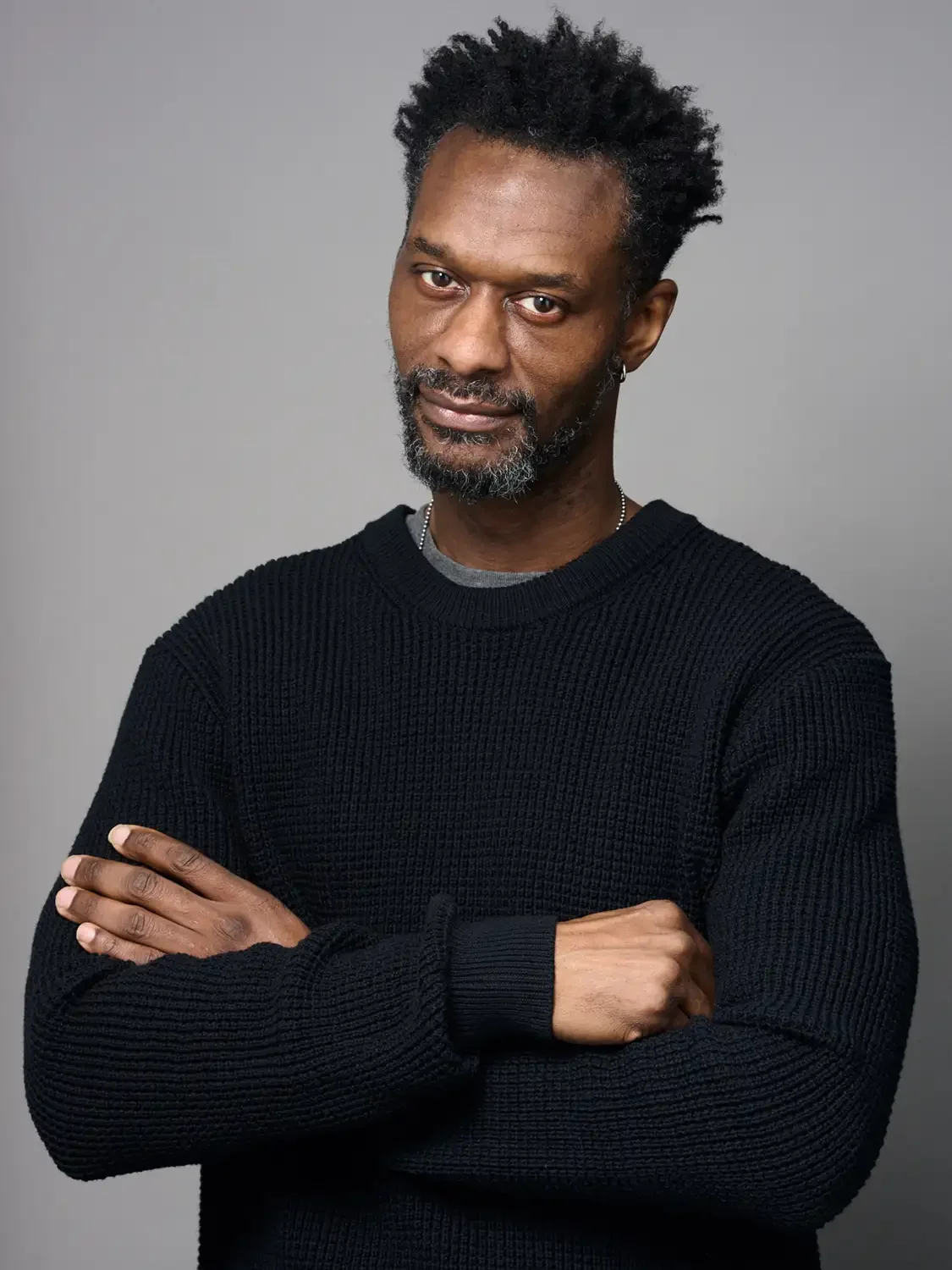 A middle-aged man with dark skin, gray beard and curly black hair, looking confident, wearing a black sweater, photographed in a professional studio