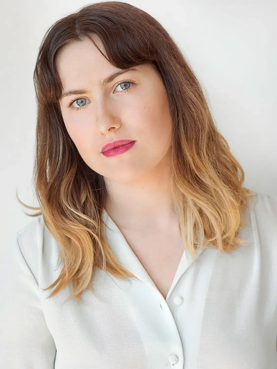 Close-up portrait of a woman with long, wavy, light brown hair with blonde tips, wearing a white collared shirt, photographed in an fashion editorial way.