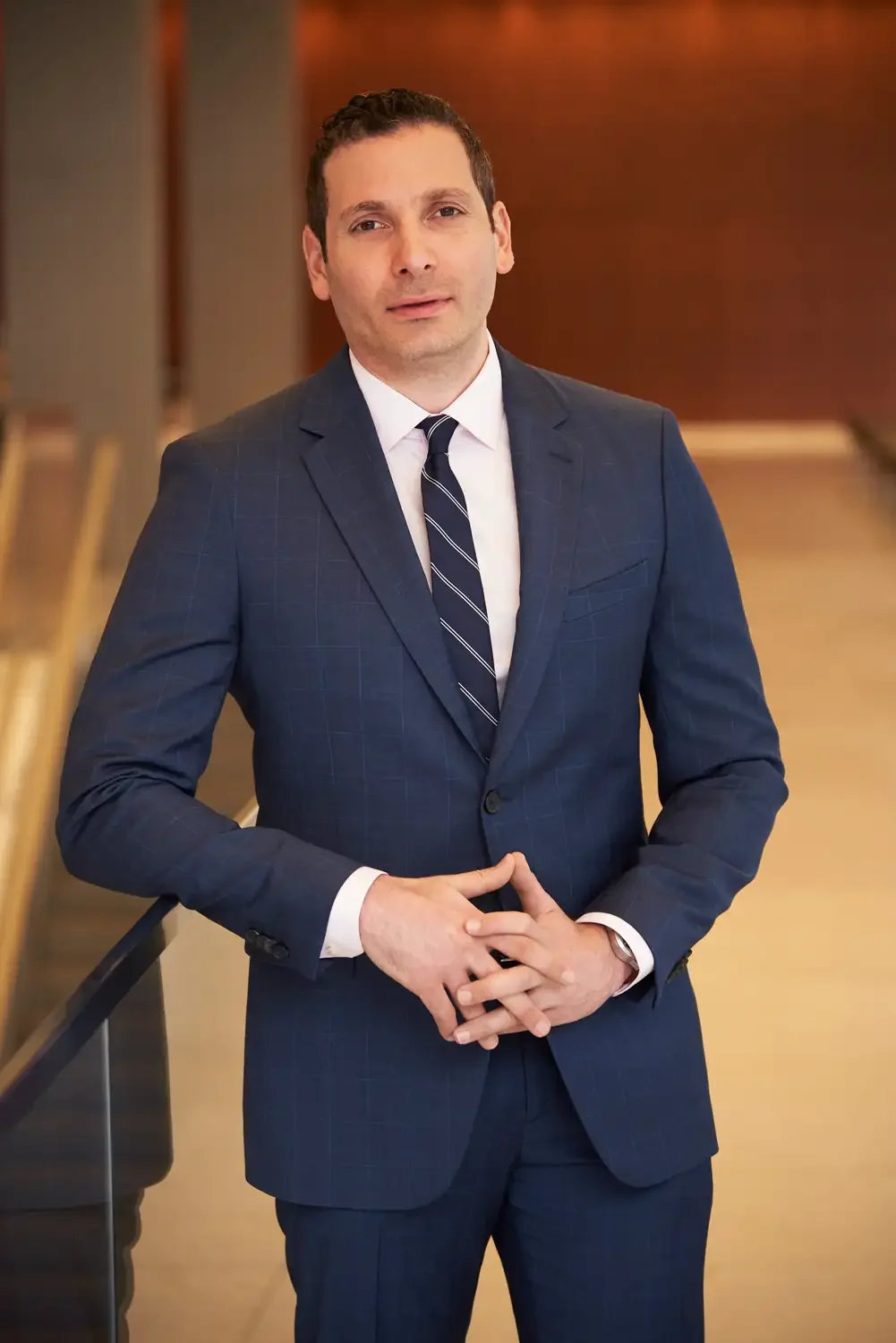 Portrait of a man in a navy blue suit with a white shirt and a dark striped tie, photographs in a corporate environment standing in a well-lit indoor setting with a wooden background.