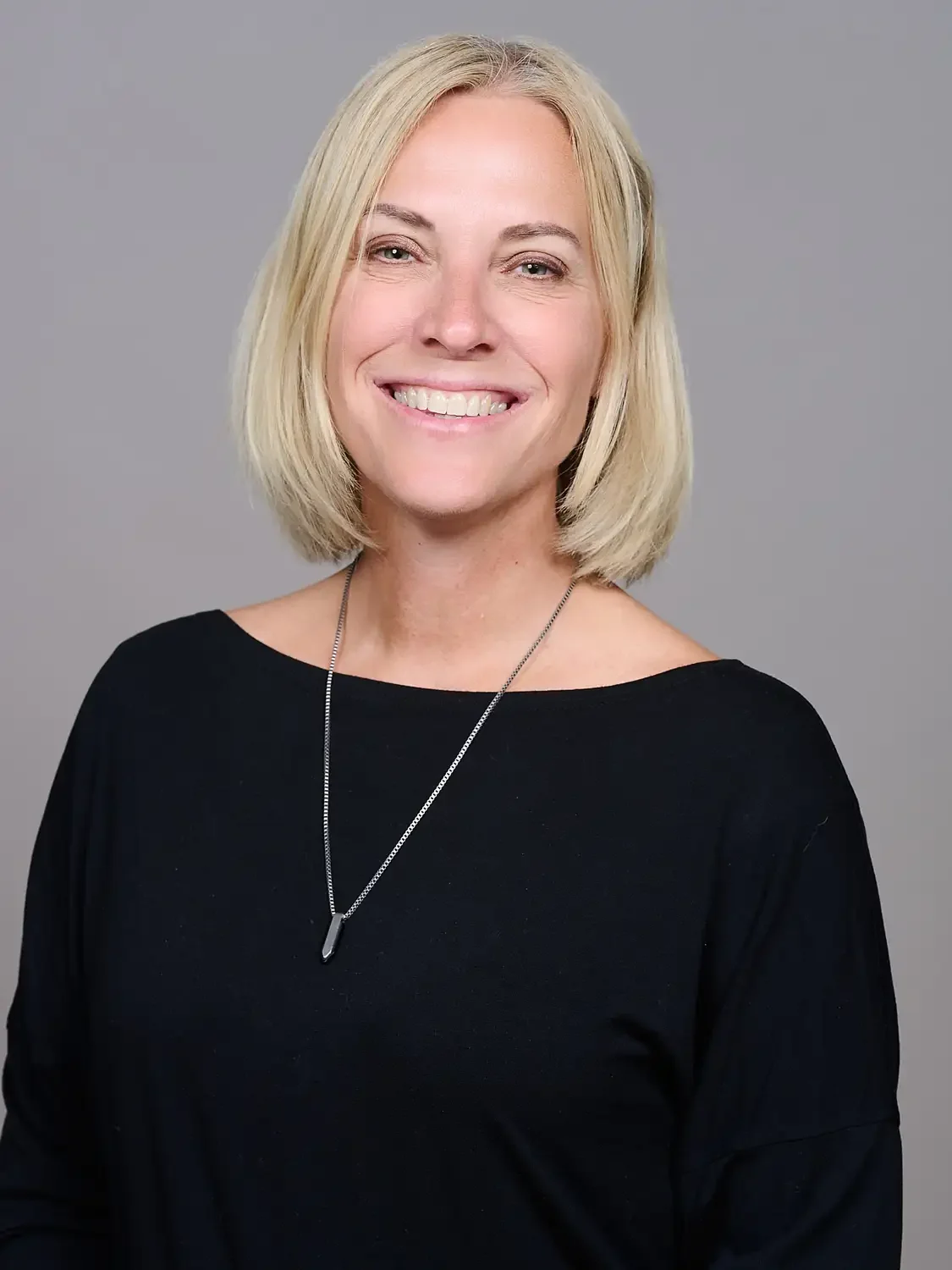 Headshot of a woman with blonde hair, smiling, wearing a black top and a silver necklace against a gray background, capturing confidence.