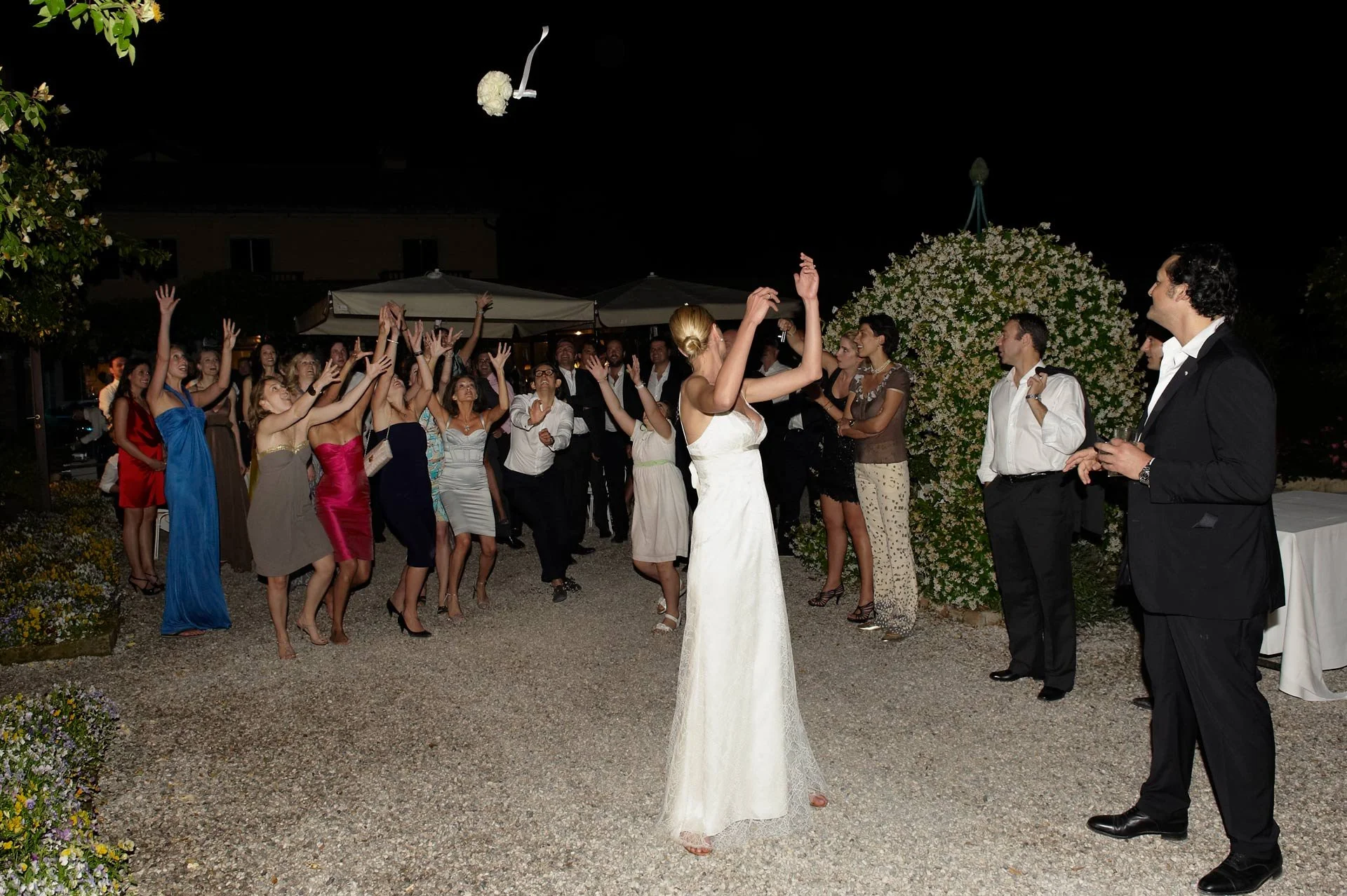 Nighttime outdoor wedding celebration with guests dancing and celebrating around bride in white dress and groom in tuxedo.