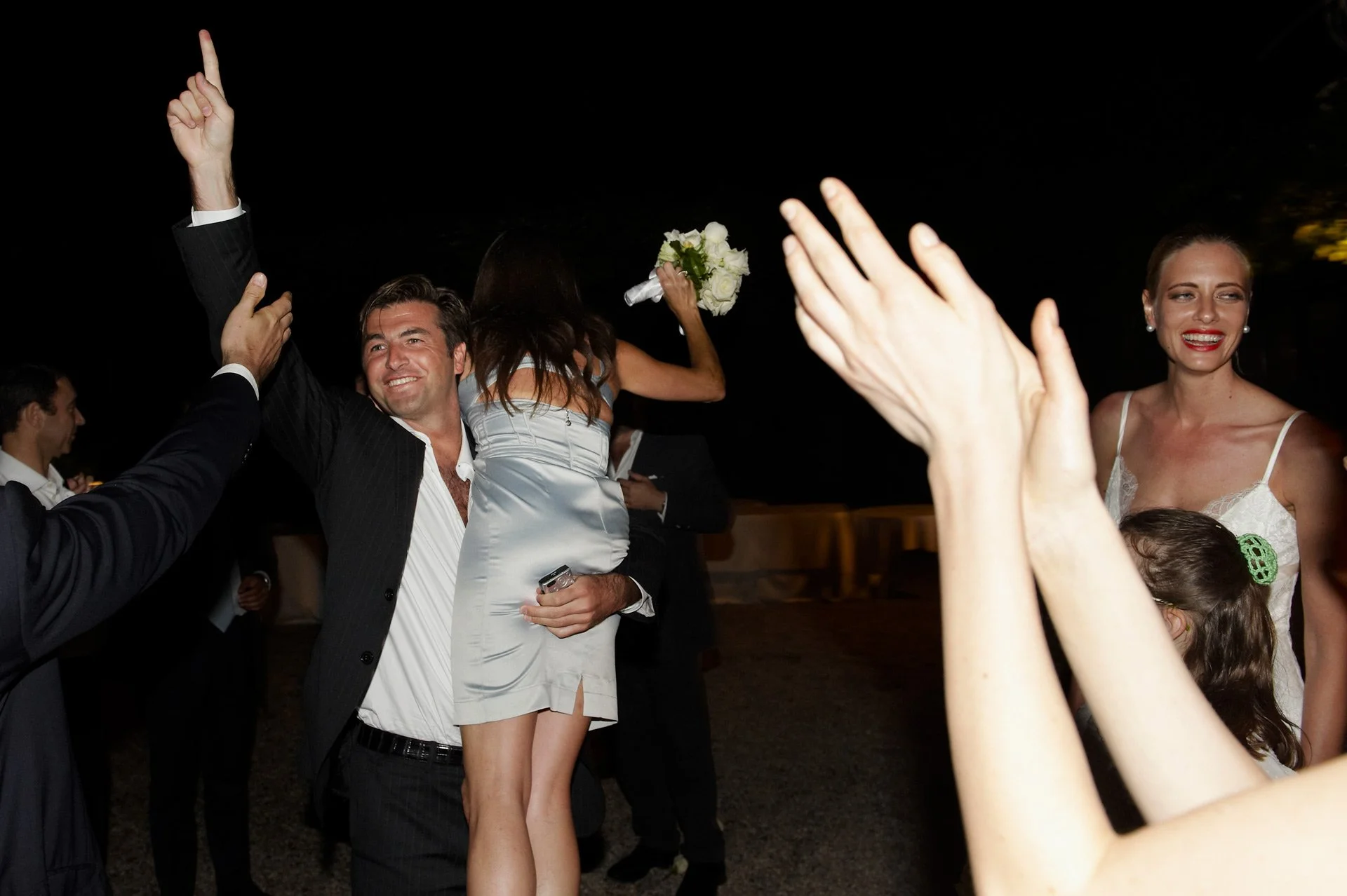 People celebrating at a wedding reception outdoors at night, with a man holding a woman with a bouquet of flowers on his shoulders, others clapping and smiling.