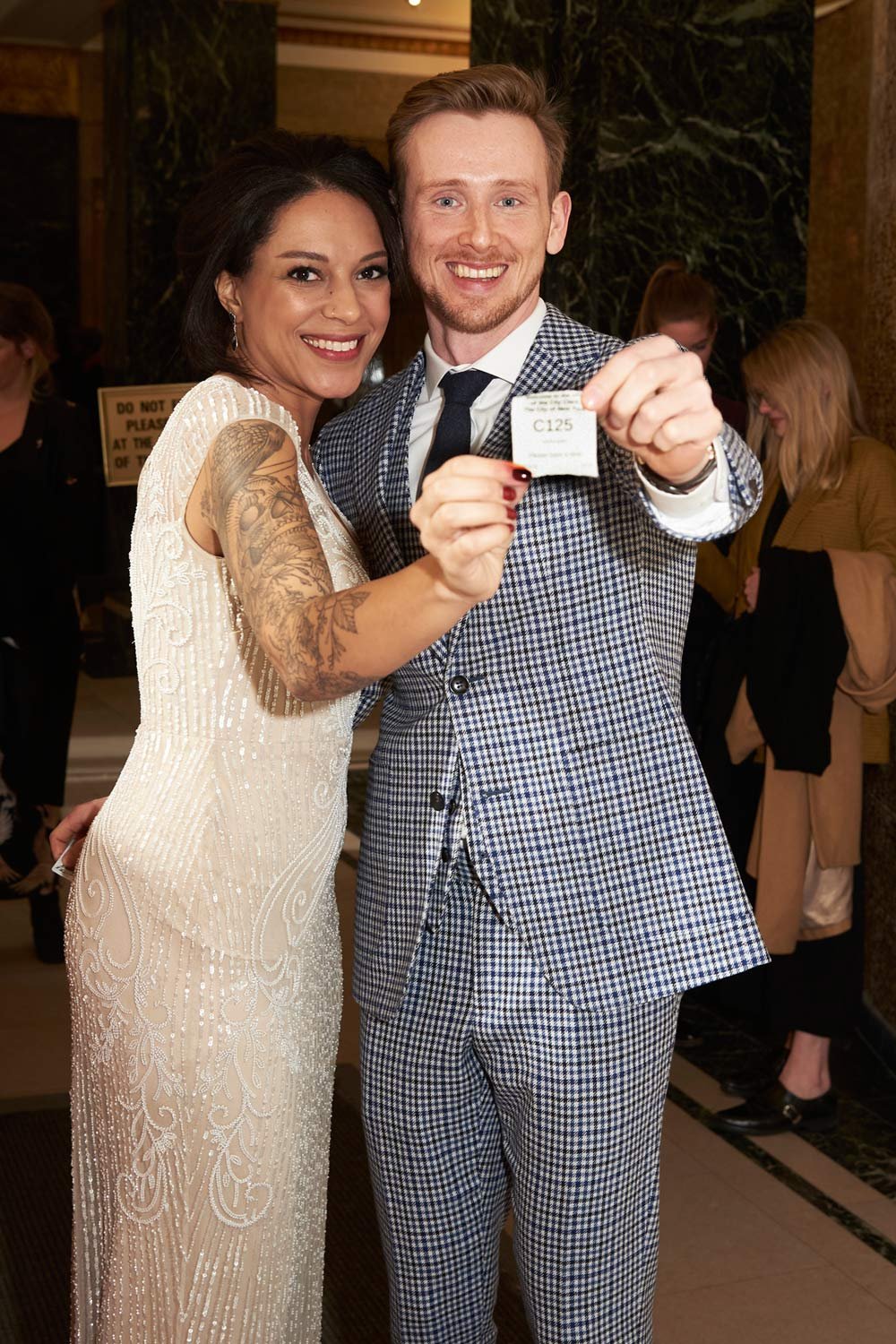 A smiling couple at a formal event holding a ticket stub. The woman has dark hair, a tattoo on her arm, and a white, beaded dress. The man has light hair, a beard, and is wearing a patterned suit with a tie.