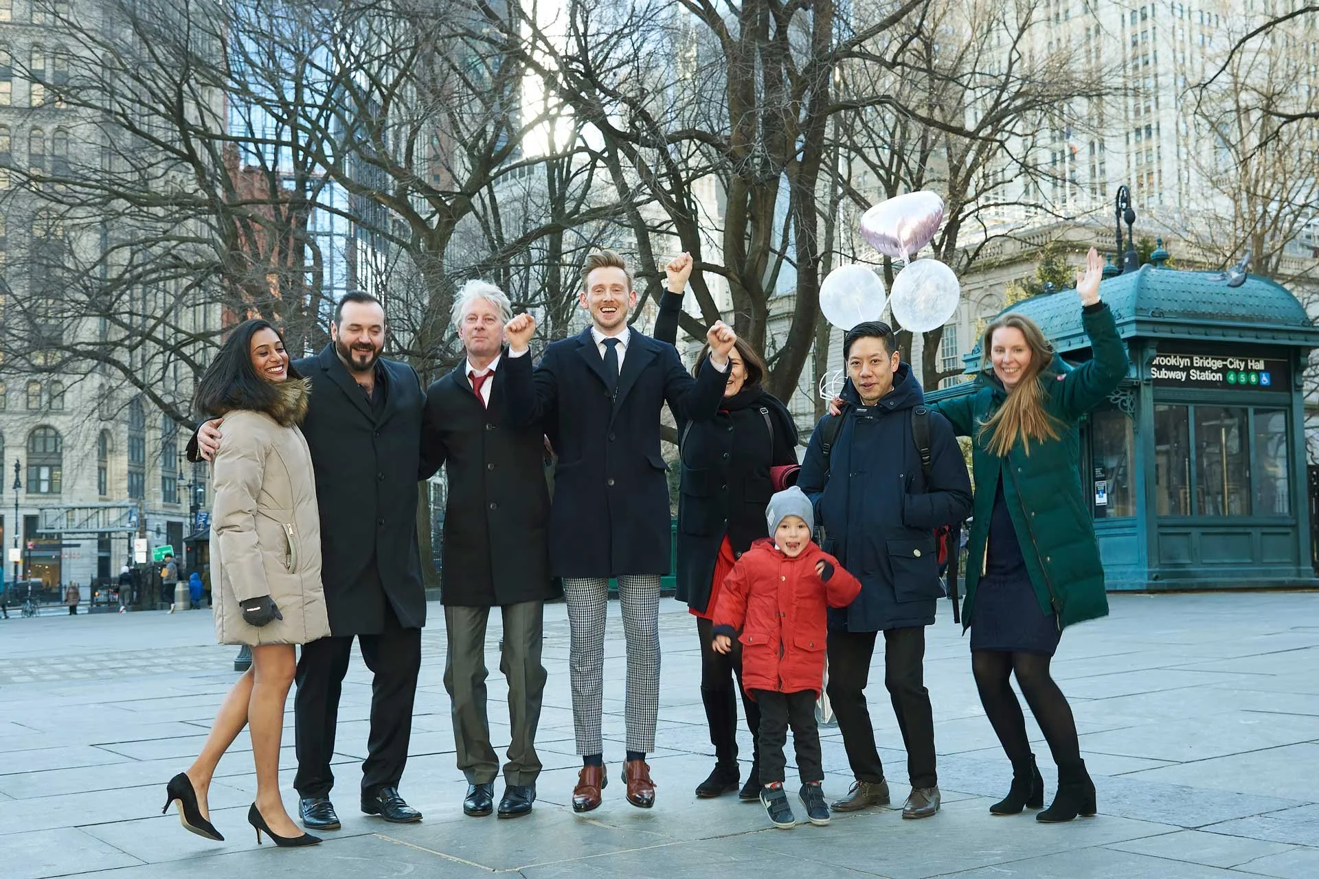 Group of smiling people celebrating outdoors in winter during daytime, with balloons, in front of a city park entrance, Brooklyn Bridge-City Hall Subway Station sign in the background.