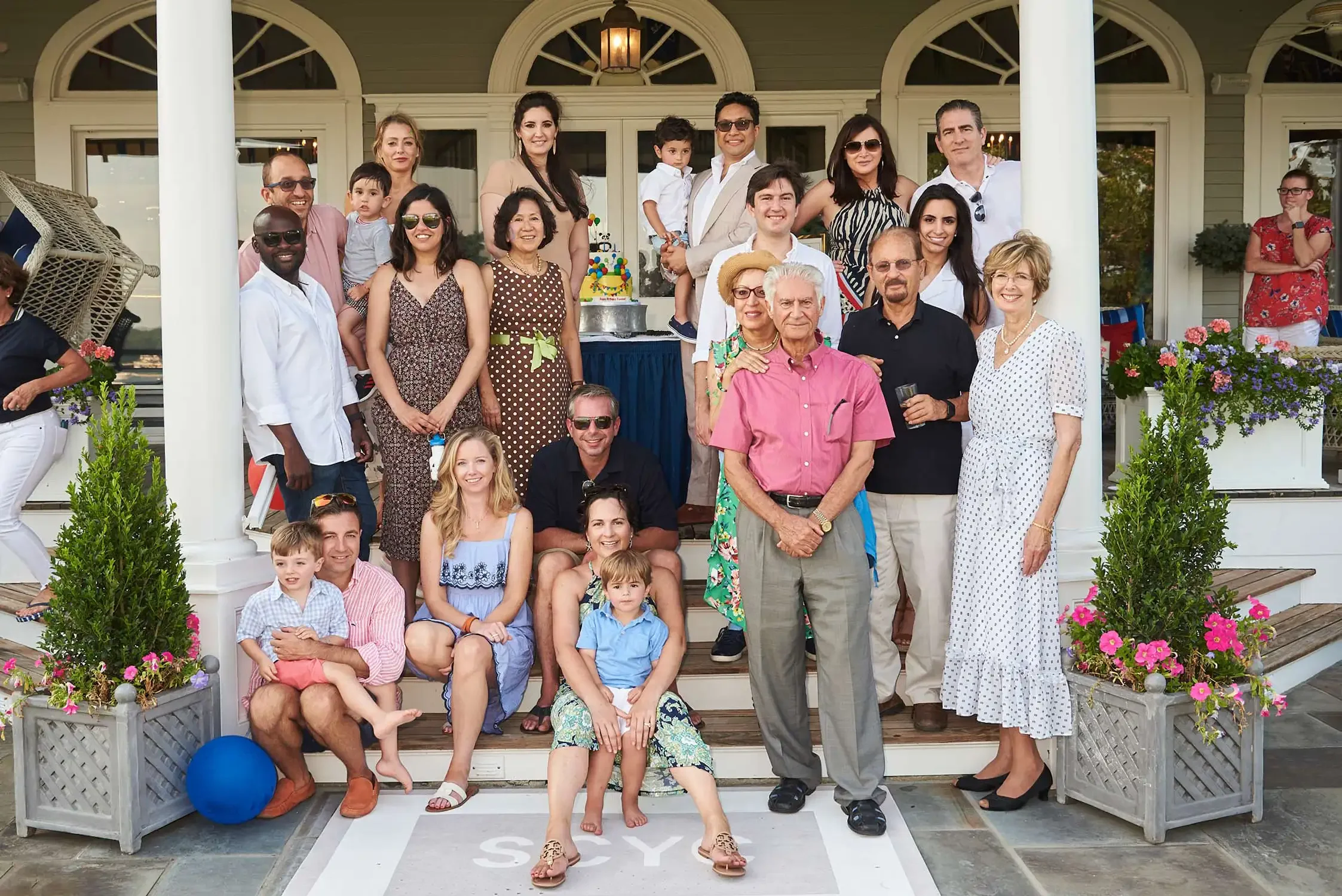 A large group of people photographed, including children and adults, gathered on the porch of a house for a celebration with a birthday cake in the center. The background shows a house with large windows and a porch with pillars.