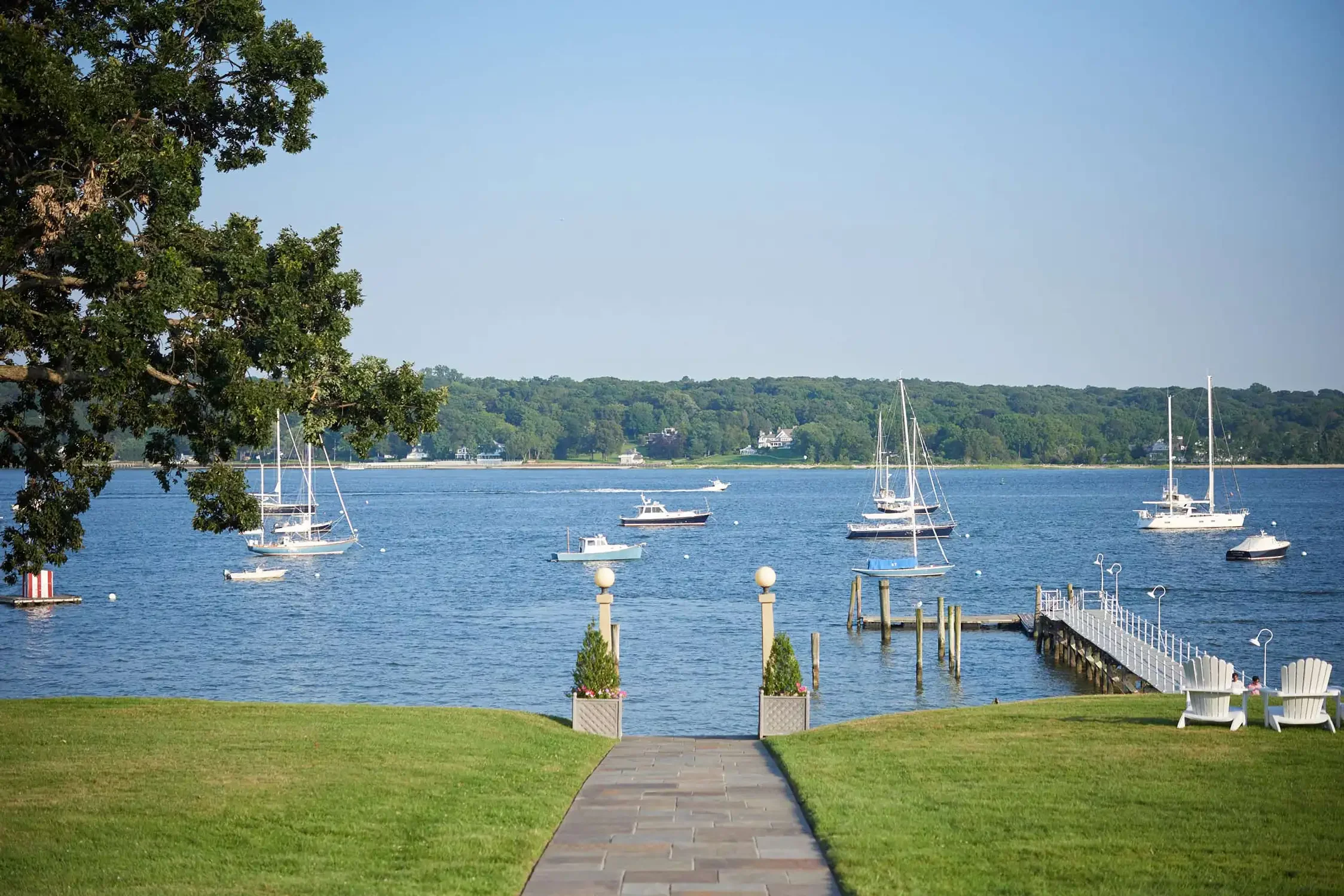 A waterfront scene with sailboats on calm water, a grassy area with a paved walkway, white chairs, and potted flowers, with trees and a hill with houses in the background under a clear blue sky.