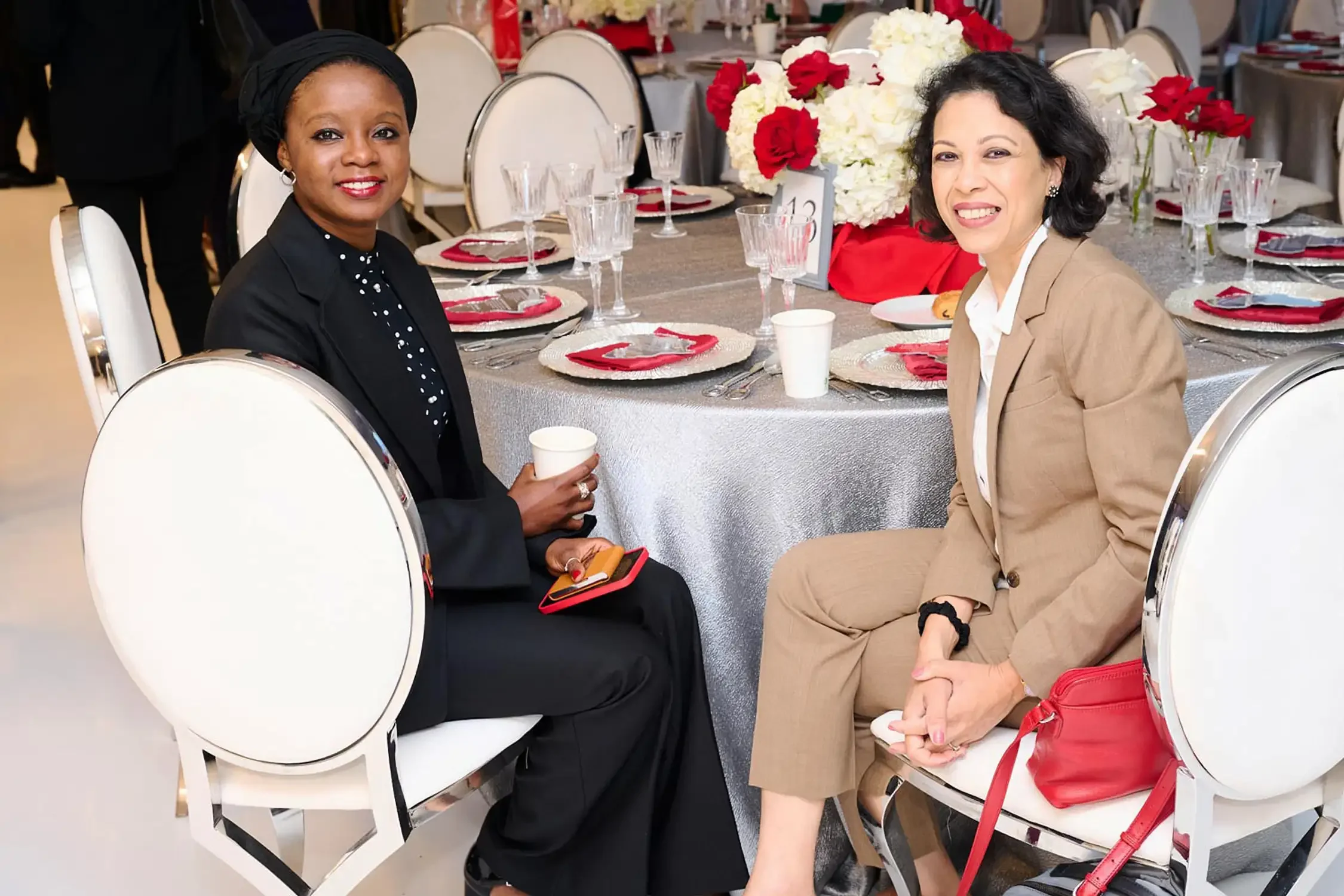 Two women, at a corporate event one African American and one Asian, sitting at a decorated banquet table with floral centerpieces, smiling and posing for the photo.
