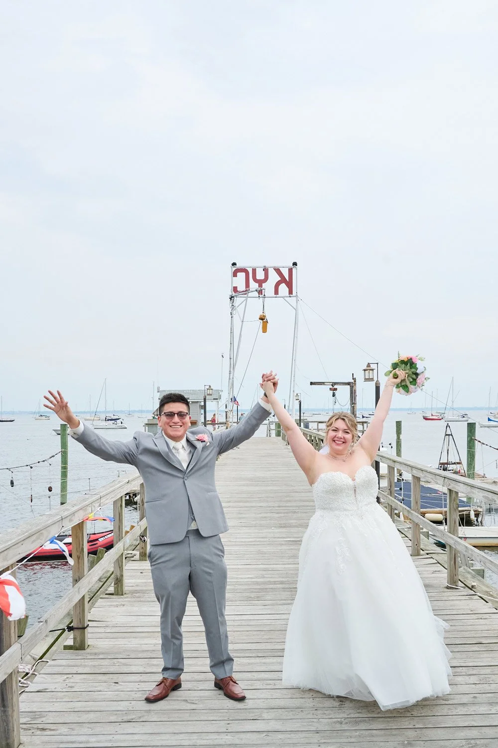 A newlywed couple celebrates happily on a wooden dock by the water, holding hands up in victory. The groom is wearing a gray suit and the bride is in a white wedding dress holding a bouquet of flowers. Sailboats are visible in the background, and an 