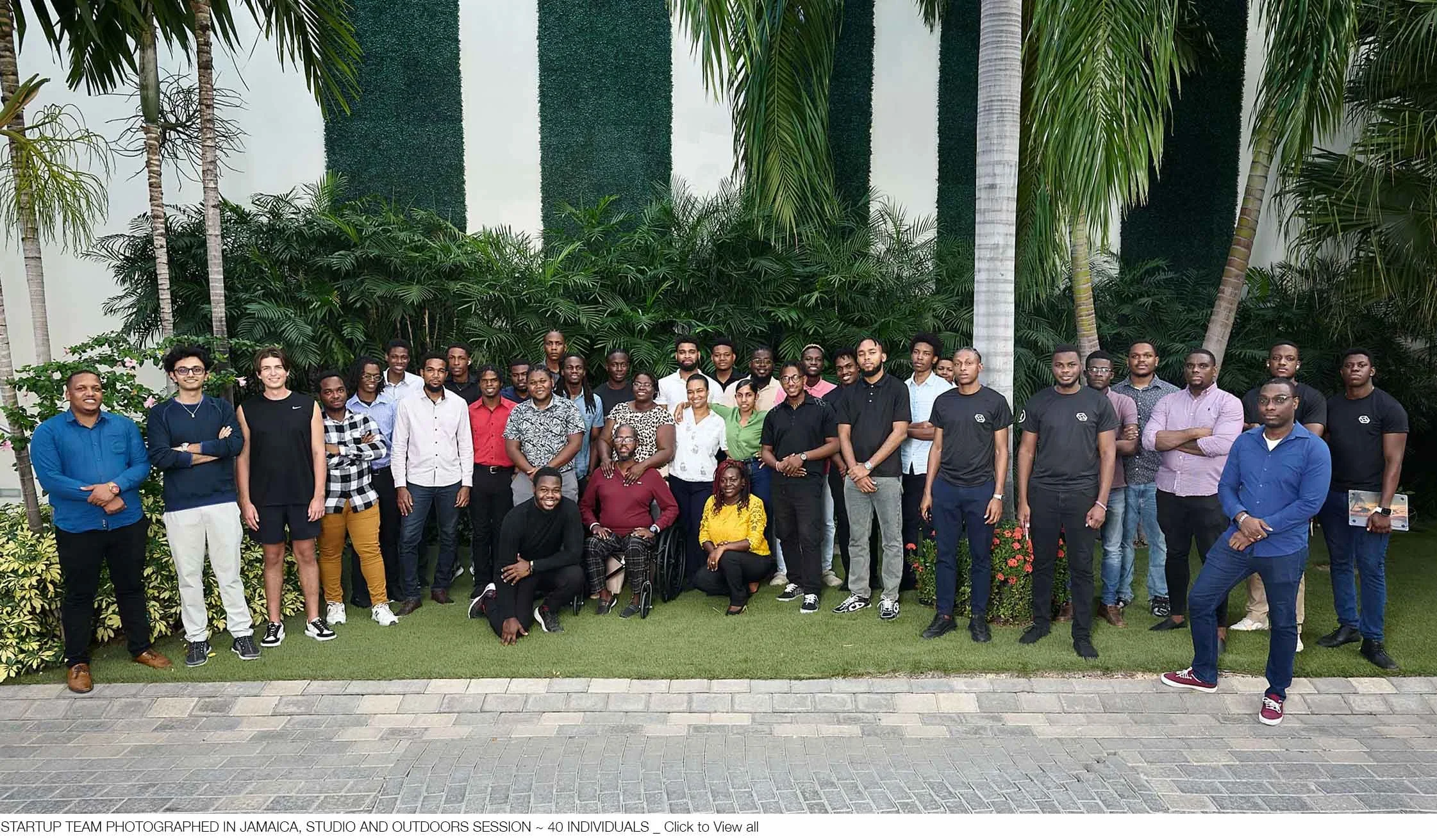 Group of 40 diverse individuals posing outdoors in front of greenery, some smiling, some with crossed arms, during a team session in Jamaica.