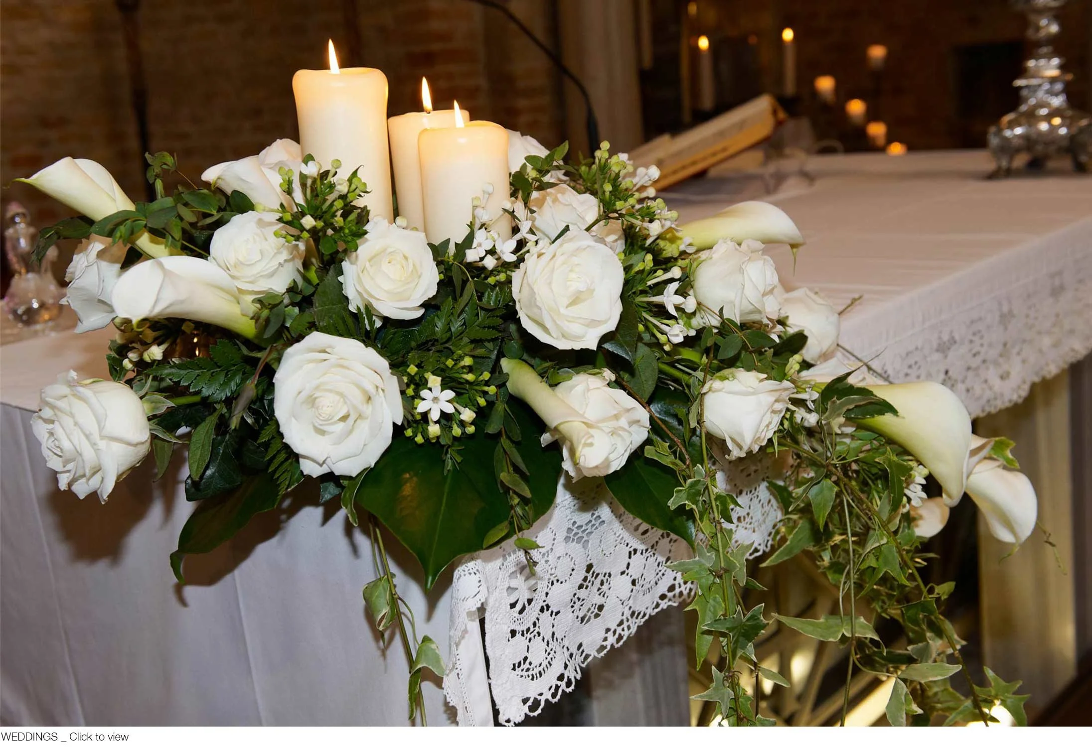 White floral arrangement with roses, calla lilies, candles, and greenery on a lace-covered table, suggesting a wedding or special event.