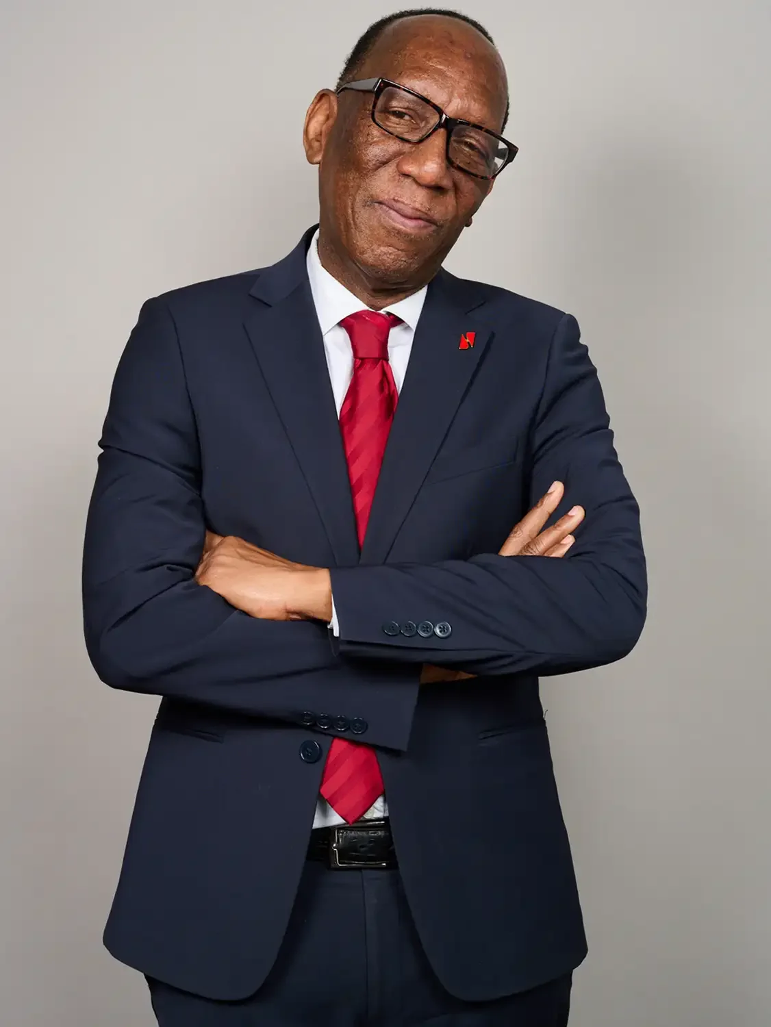 Headshot of a bank executive in a dark suit with a white shirt and red tie, wearing glasses, stands with arms crossed and a slight smile against in a photo studio