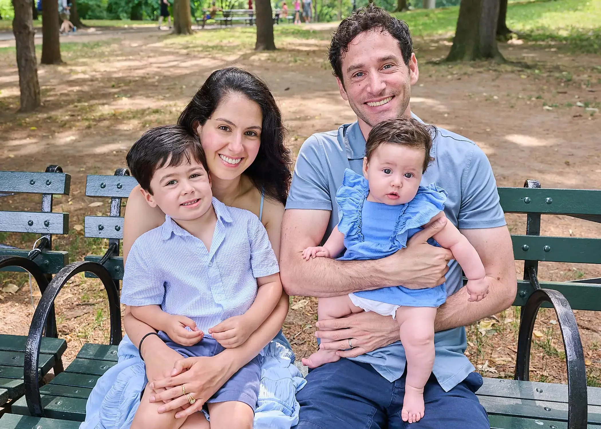 Family of four photographed sitting on a park bench, smiling, with trees and other park visitors in the background.