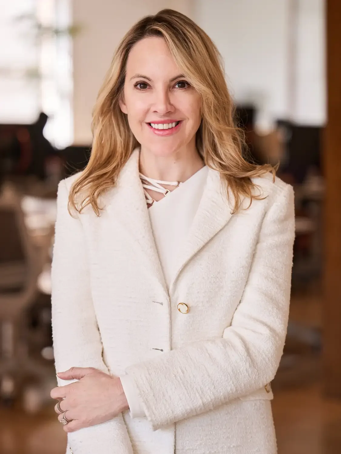 Formal headshot for an executive  woman smiling with shoulder-length blonde hair, wearing a white blazer, in a blurred office setting smiling. 