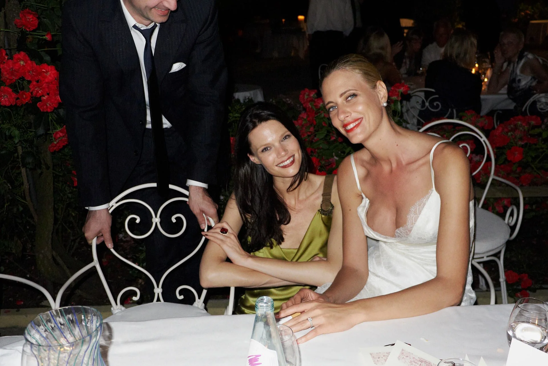 Two women sitting at a restaurant table at night, smiling. A man in a suit is standing behind them, holding a chair. The table has a glass pitcher, bottle, and glasses, with red flowers and other guests in the background.