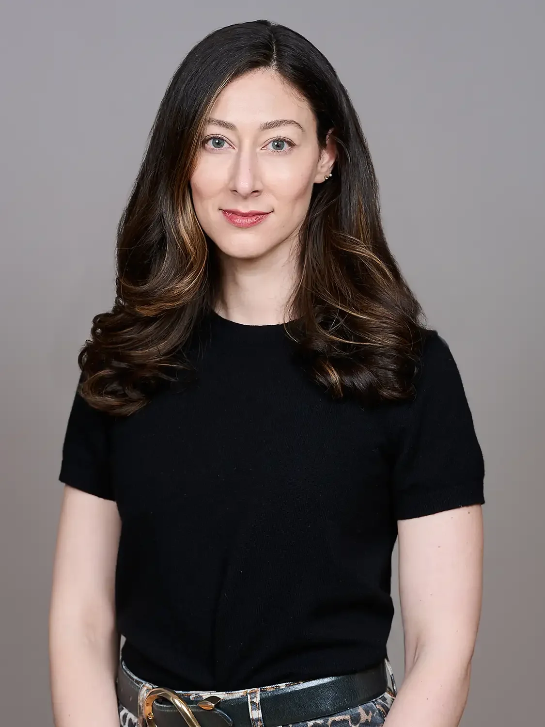 Formal portrait of a woman with long wavy brown hair wearing a black top and patterned pants photographed in a photo studio neutral grey background.