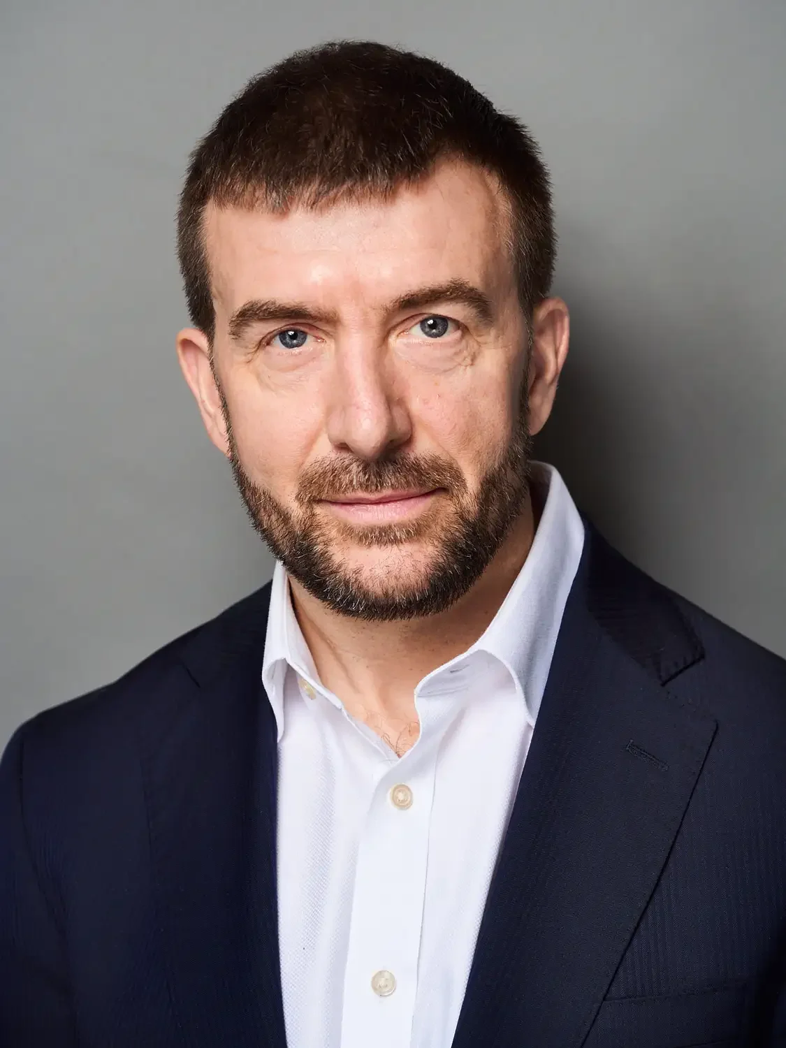 Headshot of an executive man with short brown hair, blue eyes, a beard and mustache, wearing a dark suit jacket and white dress shirt set up in a professional photo studio.