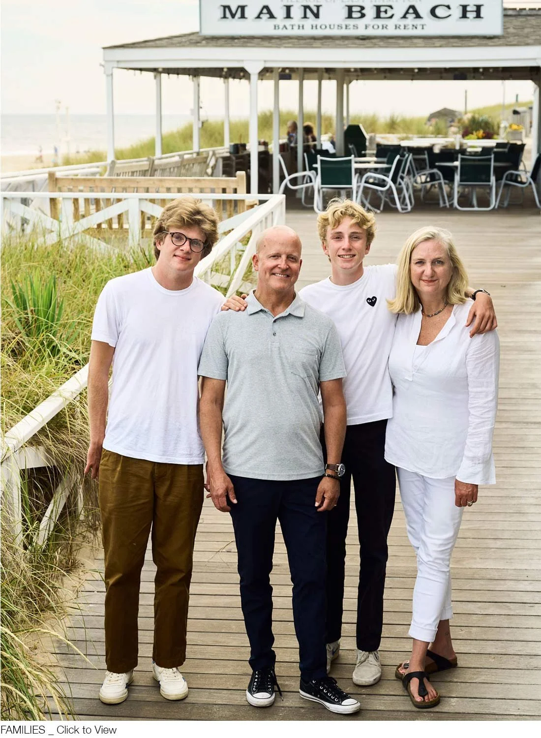 A family of four standing outdoors on a boardwalk in East Hamptons, smiling for a photo. A sign above reads 'Main Beach Bath Houses for Rent.' The family includes two young men, an older man, and an older woman.