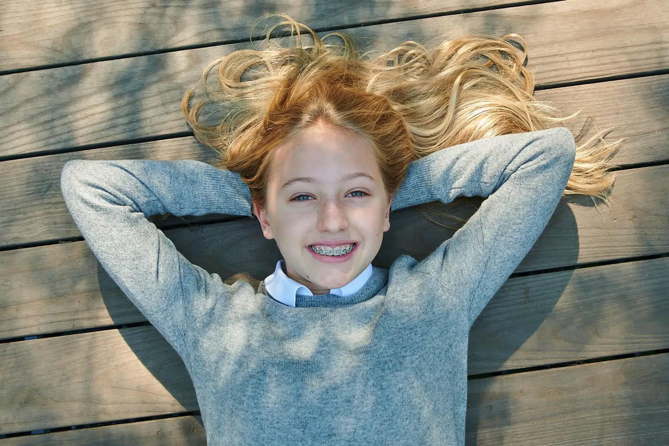 A young girl with blonde hair and braces, smiling with her eyes closed, lying on a wooden deck with her hands behind her head, wearing a grey sweater and a white shirt.