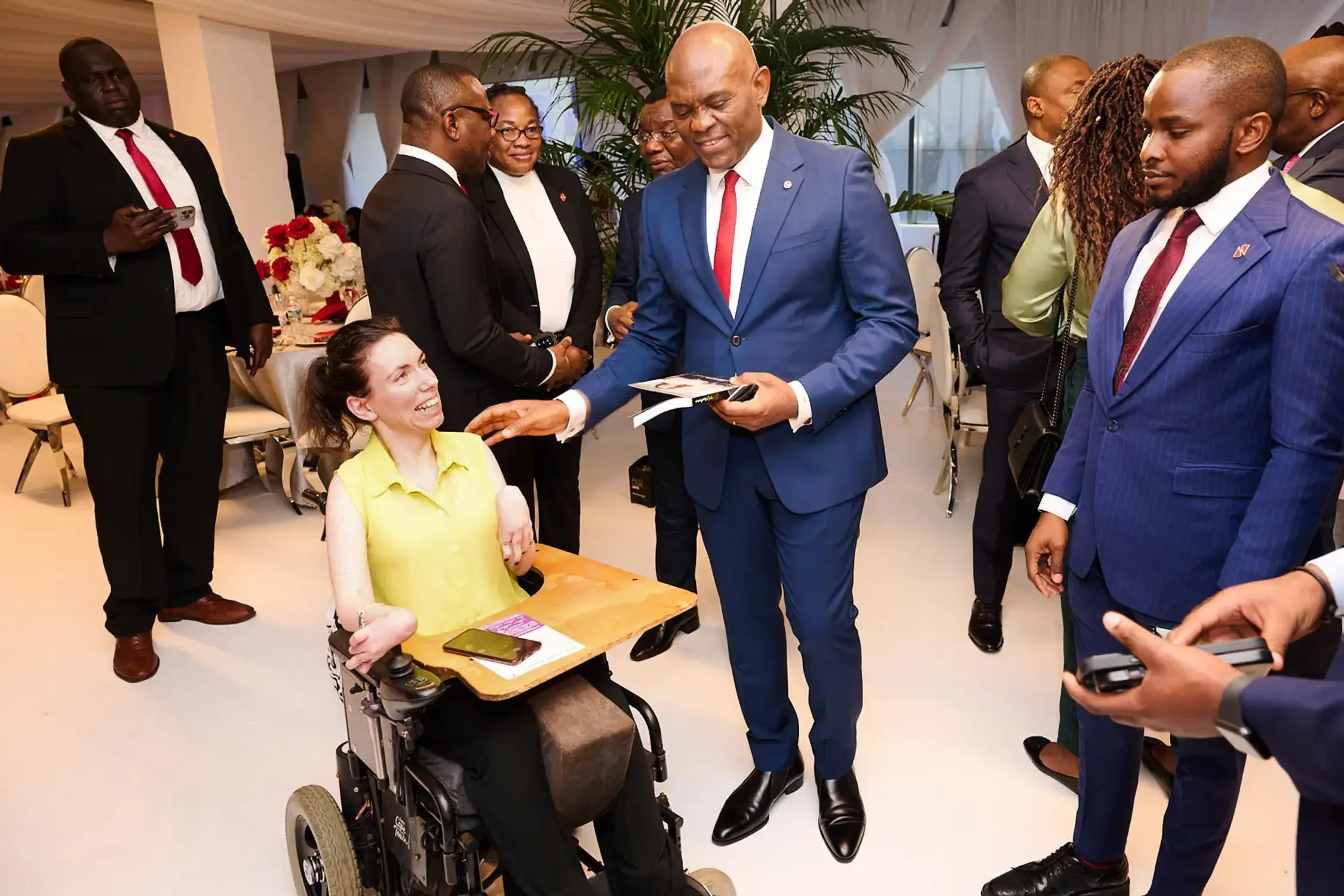 A man in a blue suit is smiling and shaking hands with a woman in a wheelchair at a corporate event with multiple other people in the background.