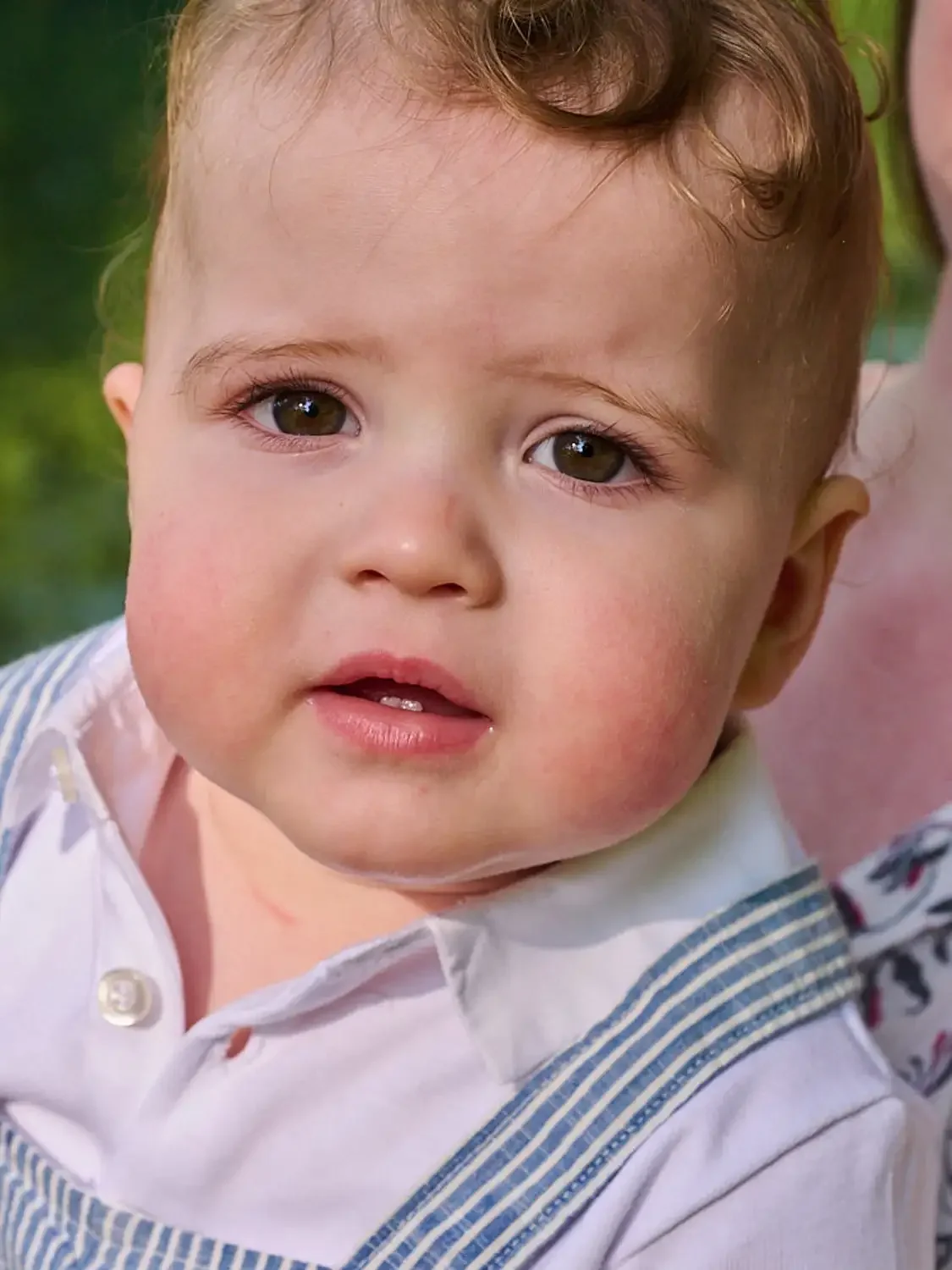 Close-up photo of a young boy with curly brown hair and brown eyes, wearing a white shirt and blue striped overalls, outdoors with a blurred green background.