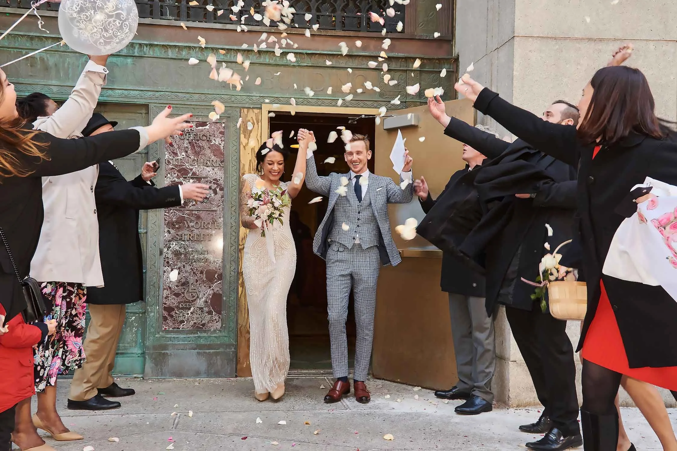 A newlywed couple exits a building amid a celebration, with friends and family throwing flower petals. The bride is holding a bouquet and wearing a lace wedding dress, and the groom is in a checkered suit. Both are smiling joyfully.