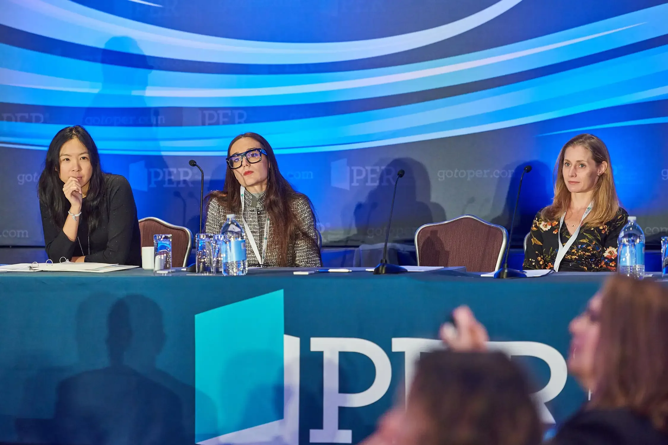Three women seated at a conference table during a panel discussion for a corporate event with a blue, wave-patterned background.