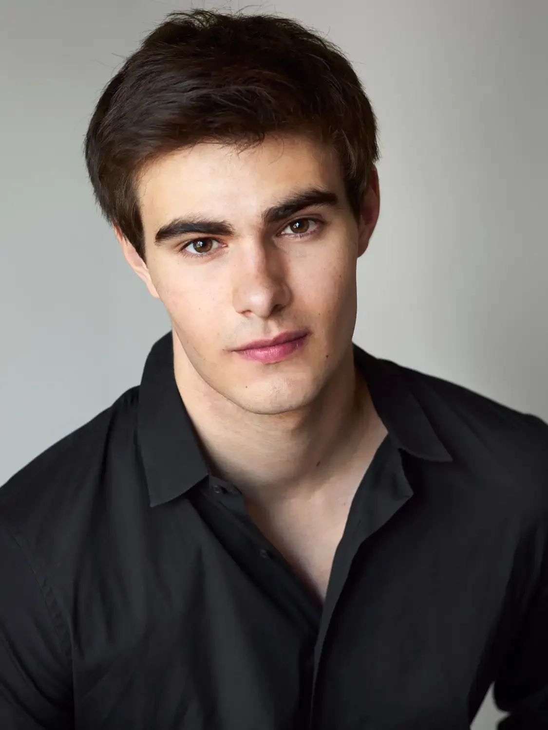 Portrait of a young man with dark brown hair, fair skin, wearing a black shirt, looking directly at the camera with a neutral expression.
