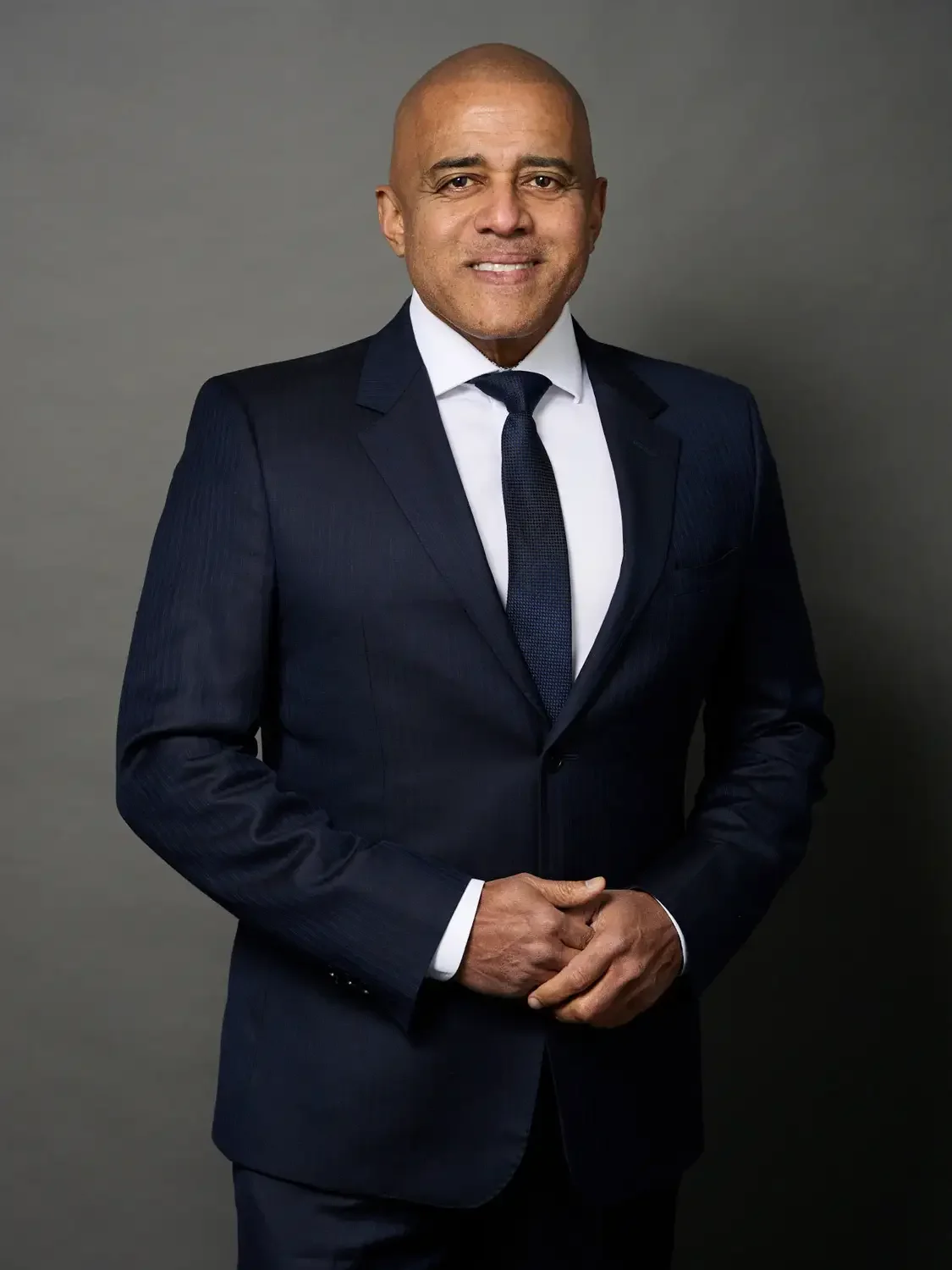 Professional portrait of a man CEO in a tailored navy suit with a shaved head, smiling with hands clasped in front. Phot studio