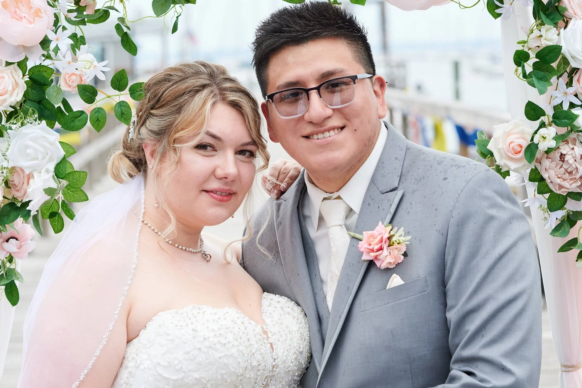 A bride and groom smiling at their wedding, standing under a floral arch with pink and white flowers and green leaves, outdoors.