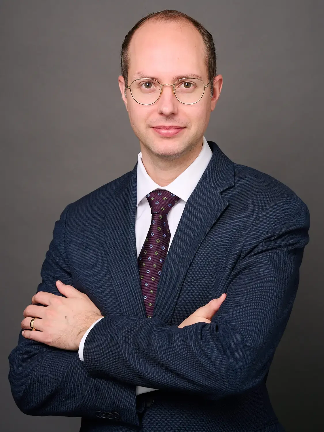Portrait of a lawyer wearing glasses, a navy suit, white shirt, and a patterned tie, standing against a gray background with his arms crossed, looking at the camera.