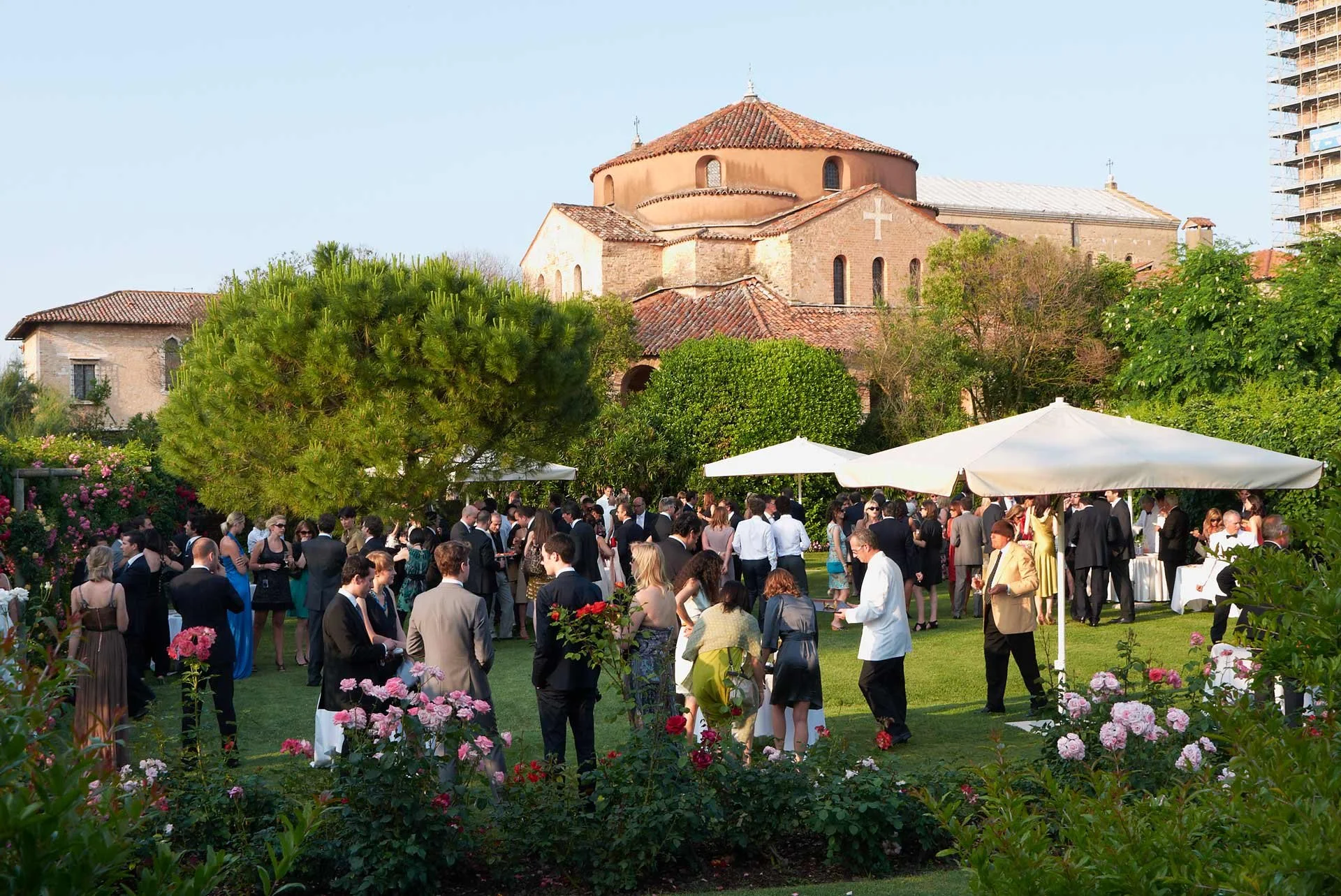 Outdoor wedding reception in Venice Italy with many guests in formal attire, white umbrellas, lush greenery, blooming flowers, and a historic brick church in the background.