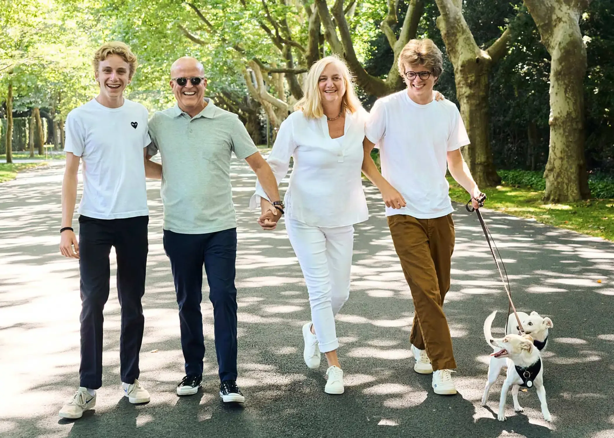 A group of five people photographed walking outdoors on a sunny day in a park lined with trees, with a woman holding hands with a man. They are smiling and appear to be enjoying each other's company.