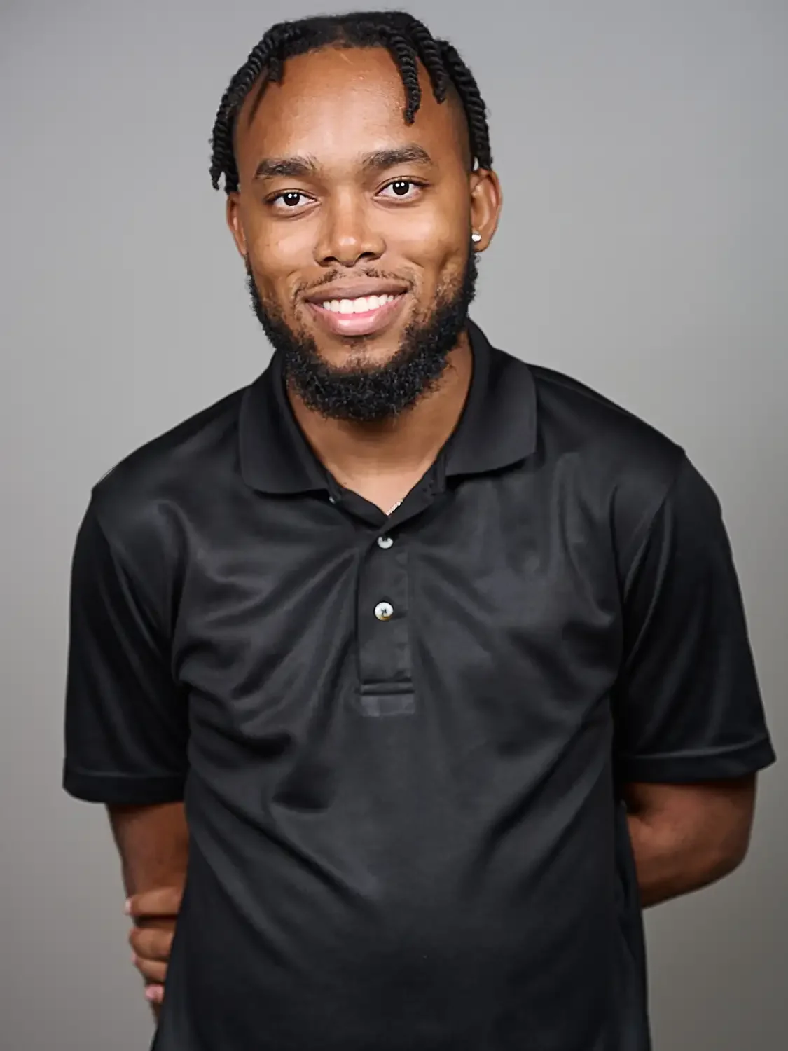 Headshot of a smiling man with dark skin and braided hair, wearing a black collared shirt, photographed in a photo studio with nice lighting.