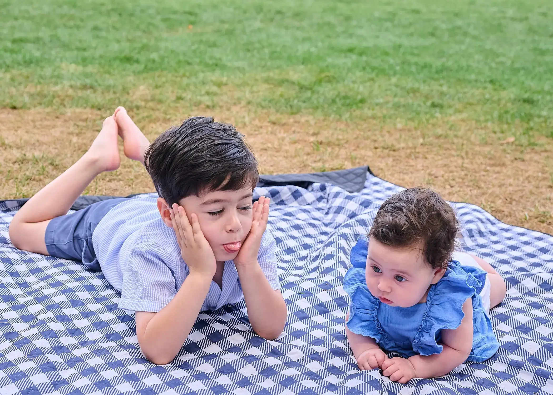 Two young childrens lying on a checkered blanket outdoors, making playful faces. One boy is resting his chin on his hands with eyes closed and tongue out.