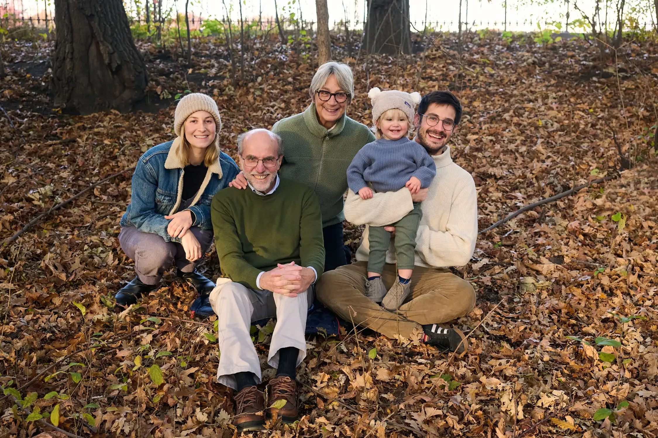 A multi-generational family of six smiling in a forest with brown fallen leaves, trees, and a fence in the background. Central Park nyc