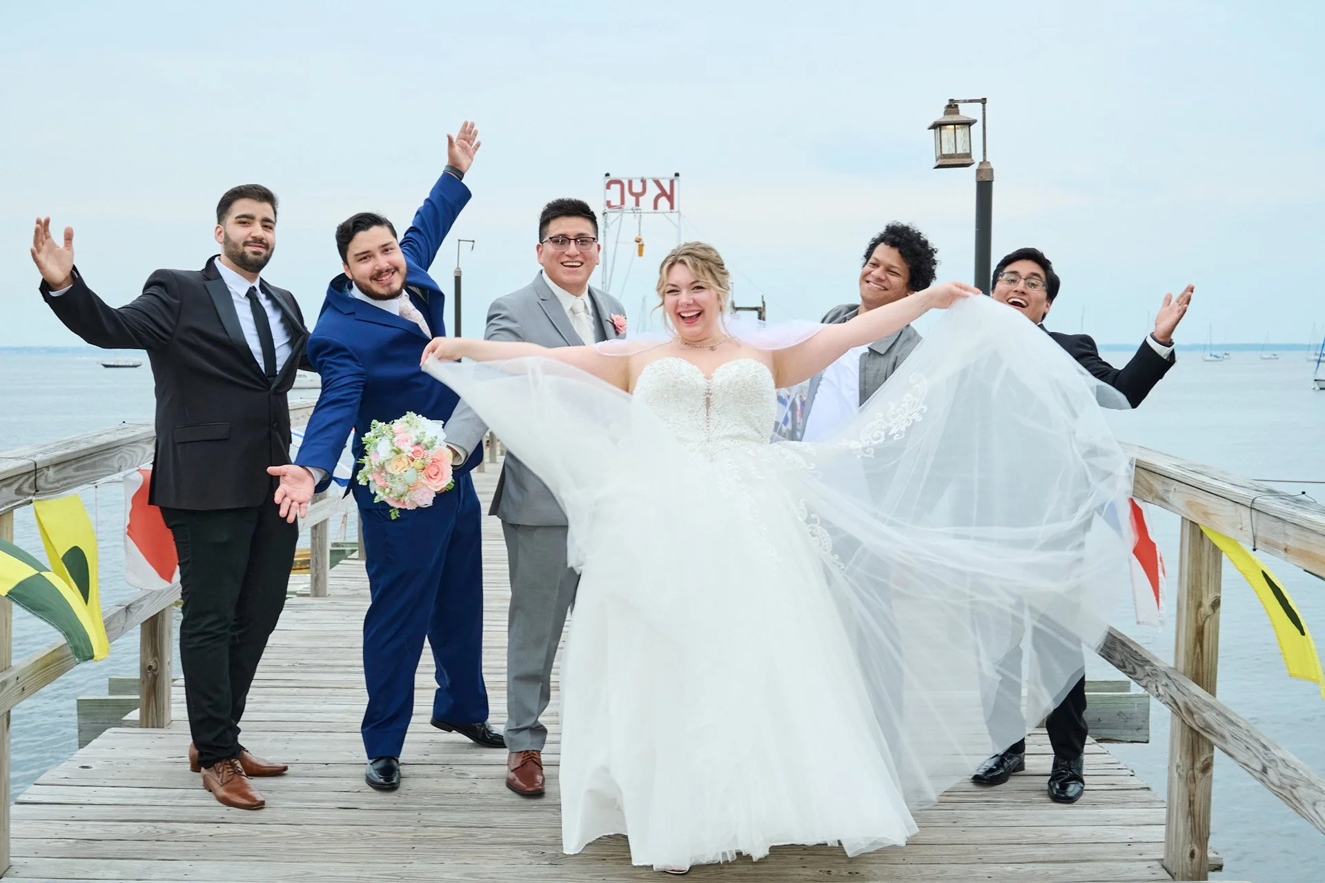 A group of six wedding guests and a bride in a white wedding dress on a wooden pier by the water. The people are smiling and celebrating, with some raising their arms. The bride is holding her dress out and standing in the center.