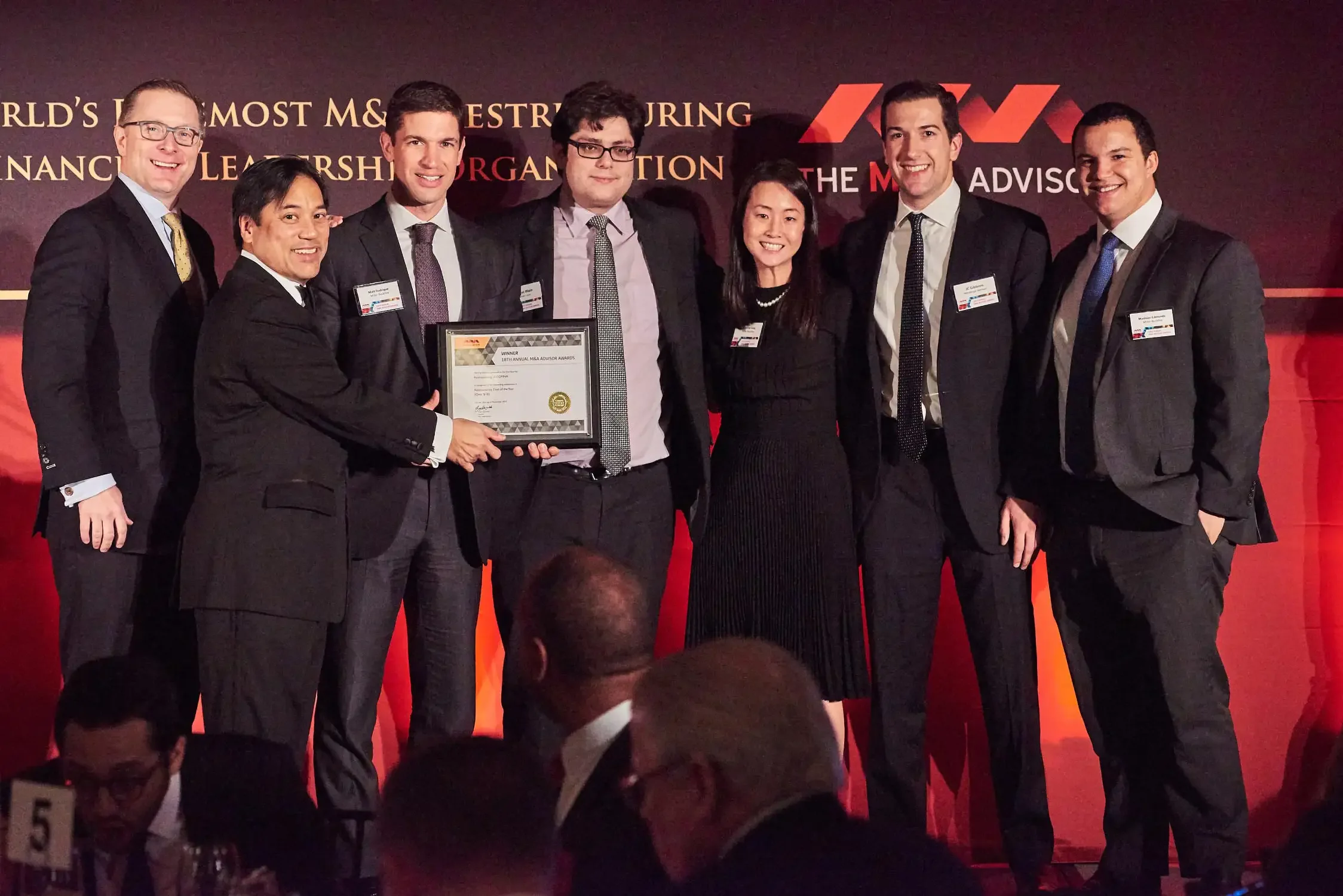 Group shot of seven people in business attire standing together on stage, they are smiling and posing for a photo.