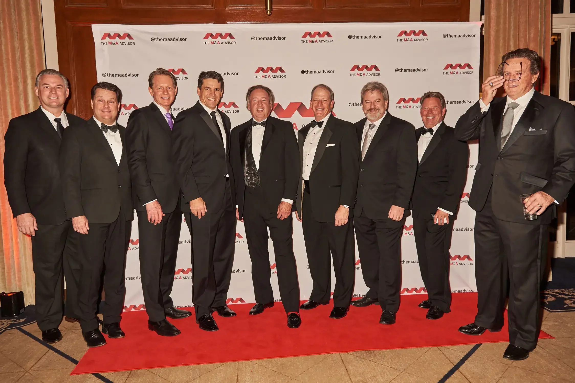 Photo of a group of men in tuxedos standing on a red carpet, celebrating at a formal event with a branded backdrop in the background.