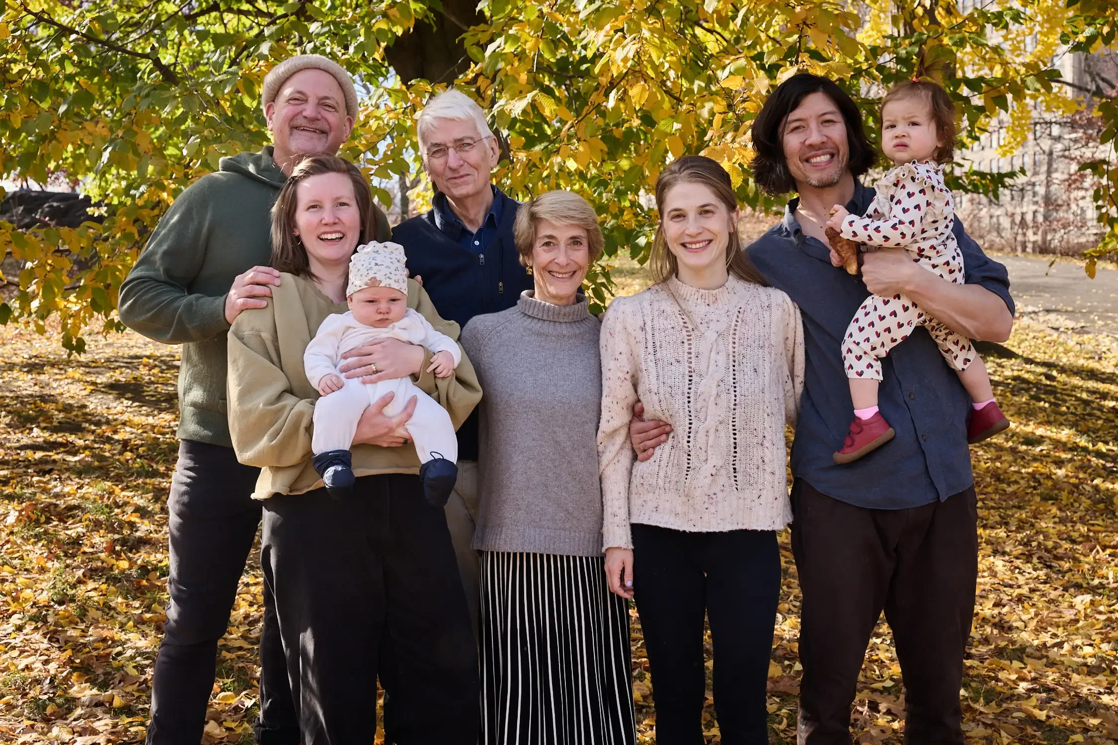 A diverse group of eight people including elderly, middle-aged, young adults, and children, photographed standing outdoors under a yellowing tree in autumn.