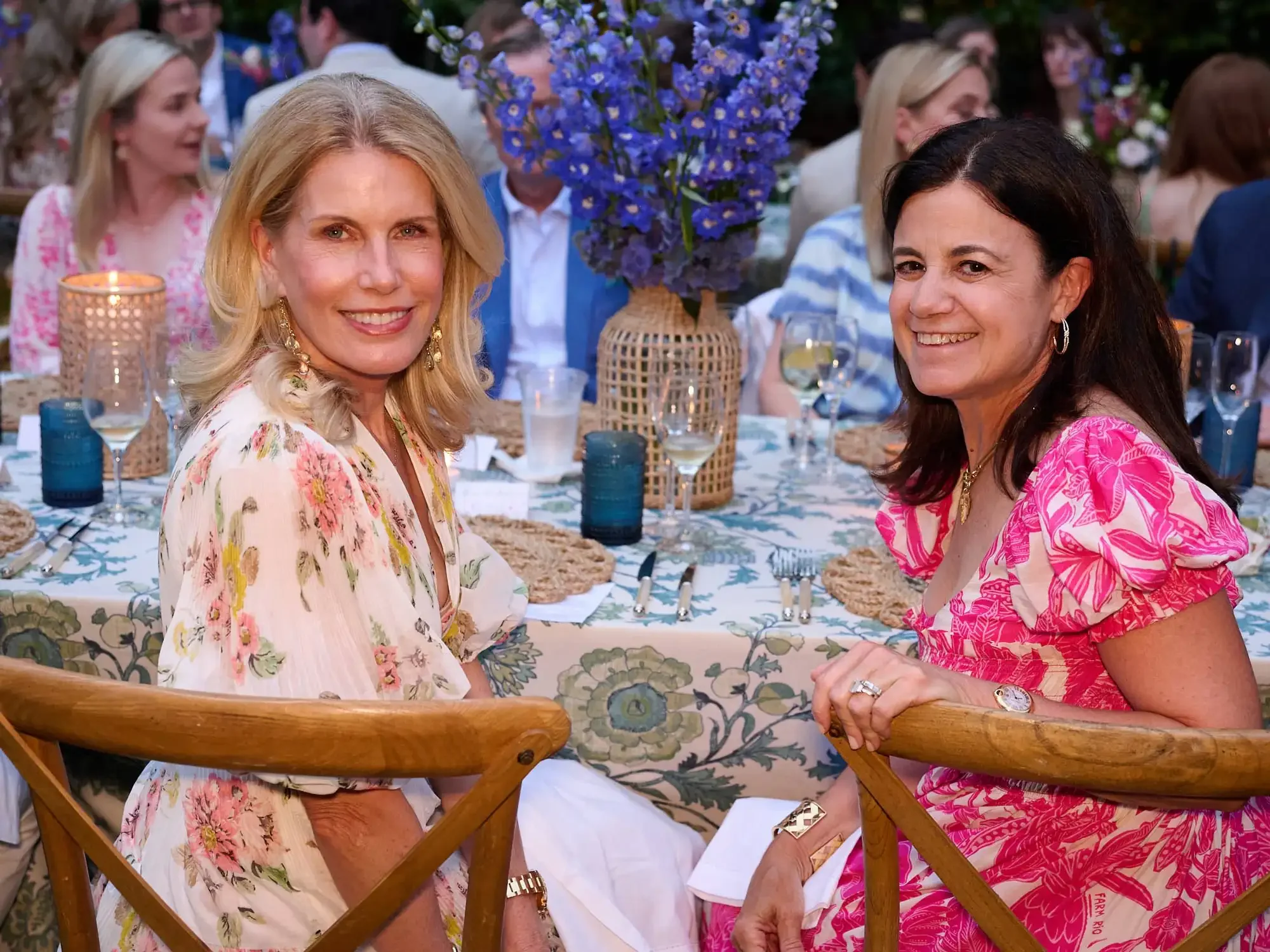 Two women during a gathering or celebration, smiling at the camera. One woman has blonde hair, wears a floral dress, and the other has dark hair, wears a pink patterned dress photographed in a luxurious setup.