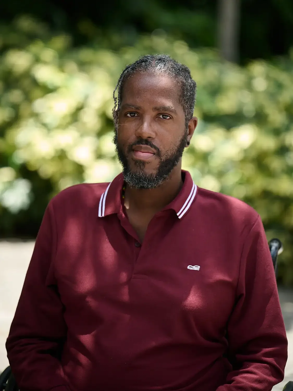 Professional headshot of a man with a beard and mustache wearing a maroon polo shirt with white stripes on the collar, sitting outdoors with green foliage in the background.