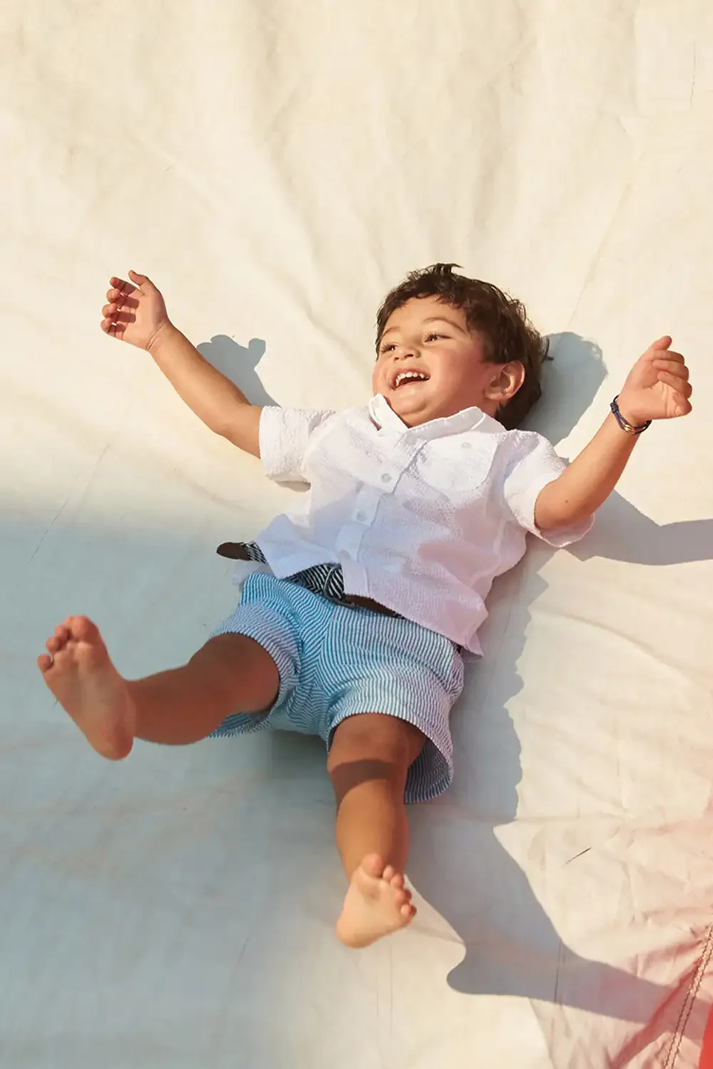 Family photographs of a young boy lying on a white surface, smiling with arms spread out, wearing a white shirt and blue striped shorts.
