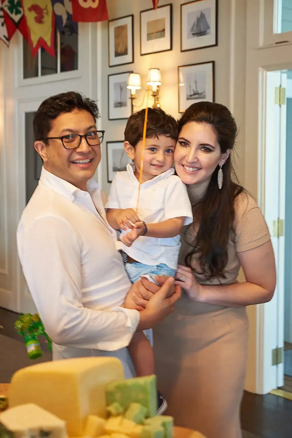 Family photos of three smiling and celebrating indoors, with colorful banners hanging on the wall and framed artwork in the background. The father holds a young birthday  boy