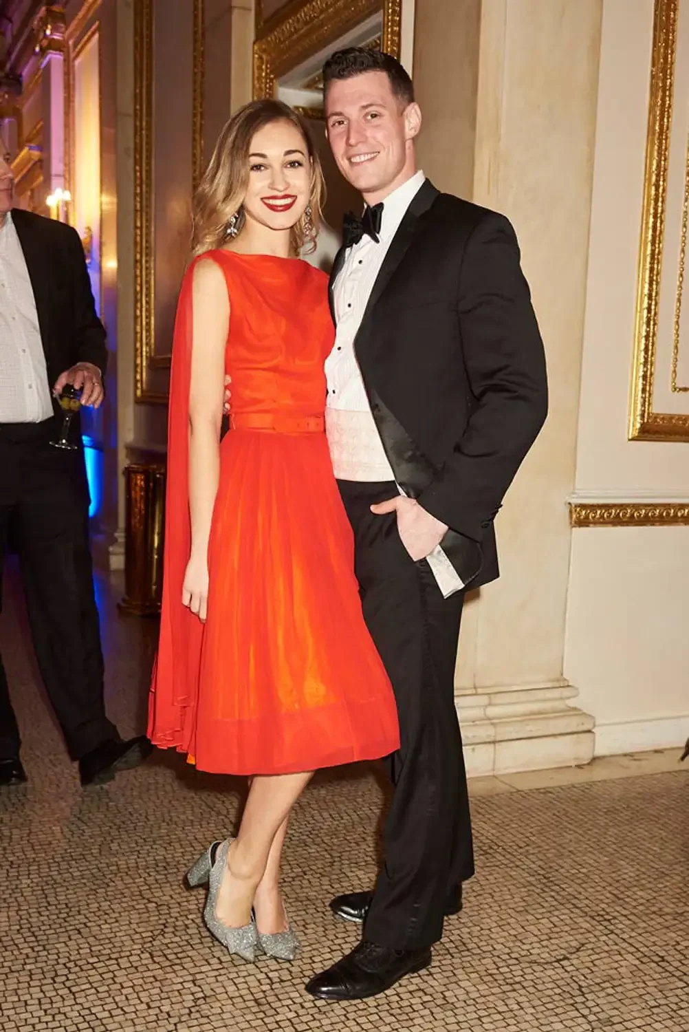 A couple dressed in formal attire, standing together at a corporate event. They are standing in a richly decorated room with gold-framed mirrors.